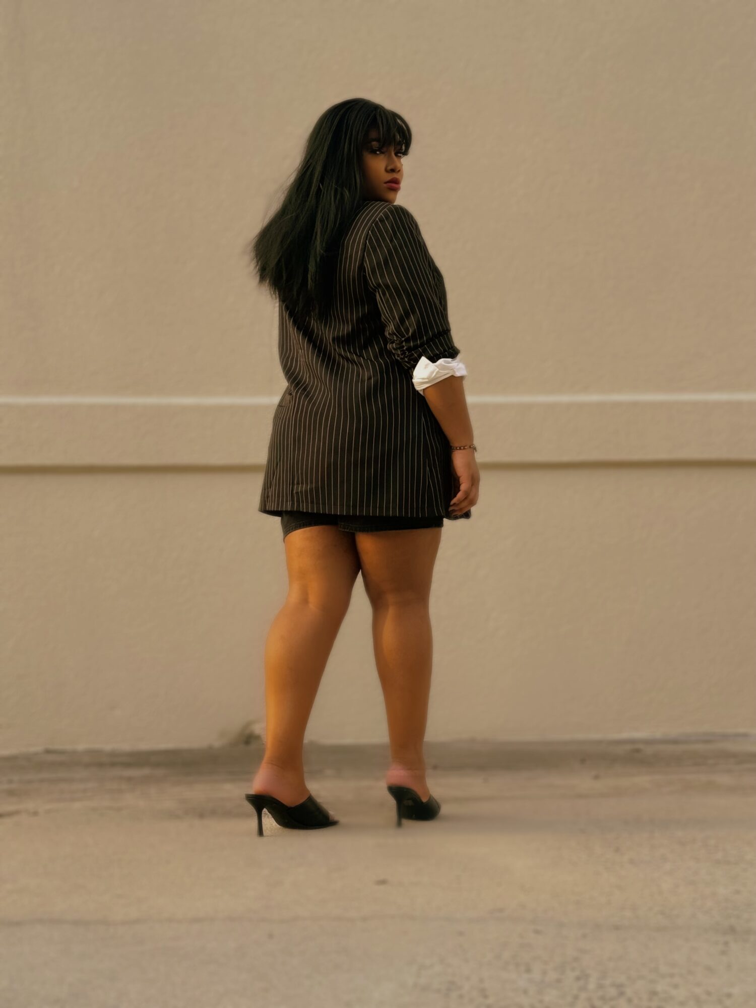 Woman standing sideways in a pinstripe blazer with rolled-up sleeves, wearing high heels, against a plain wall.