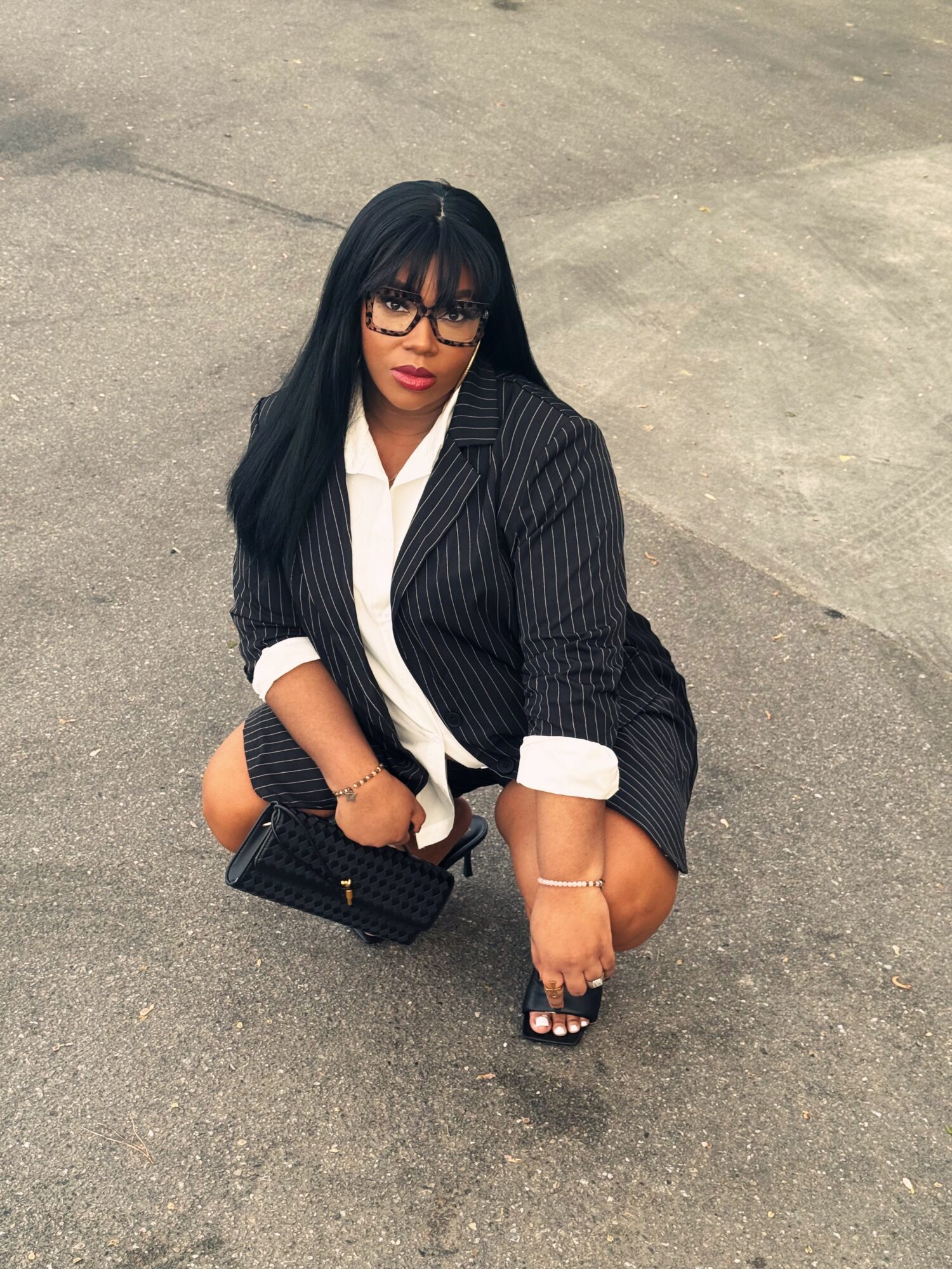 Woman crouching on pavement, wearing glasses, a pinstripe blazer, and holding a black purse.