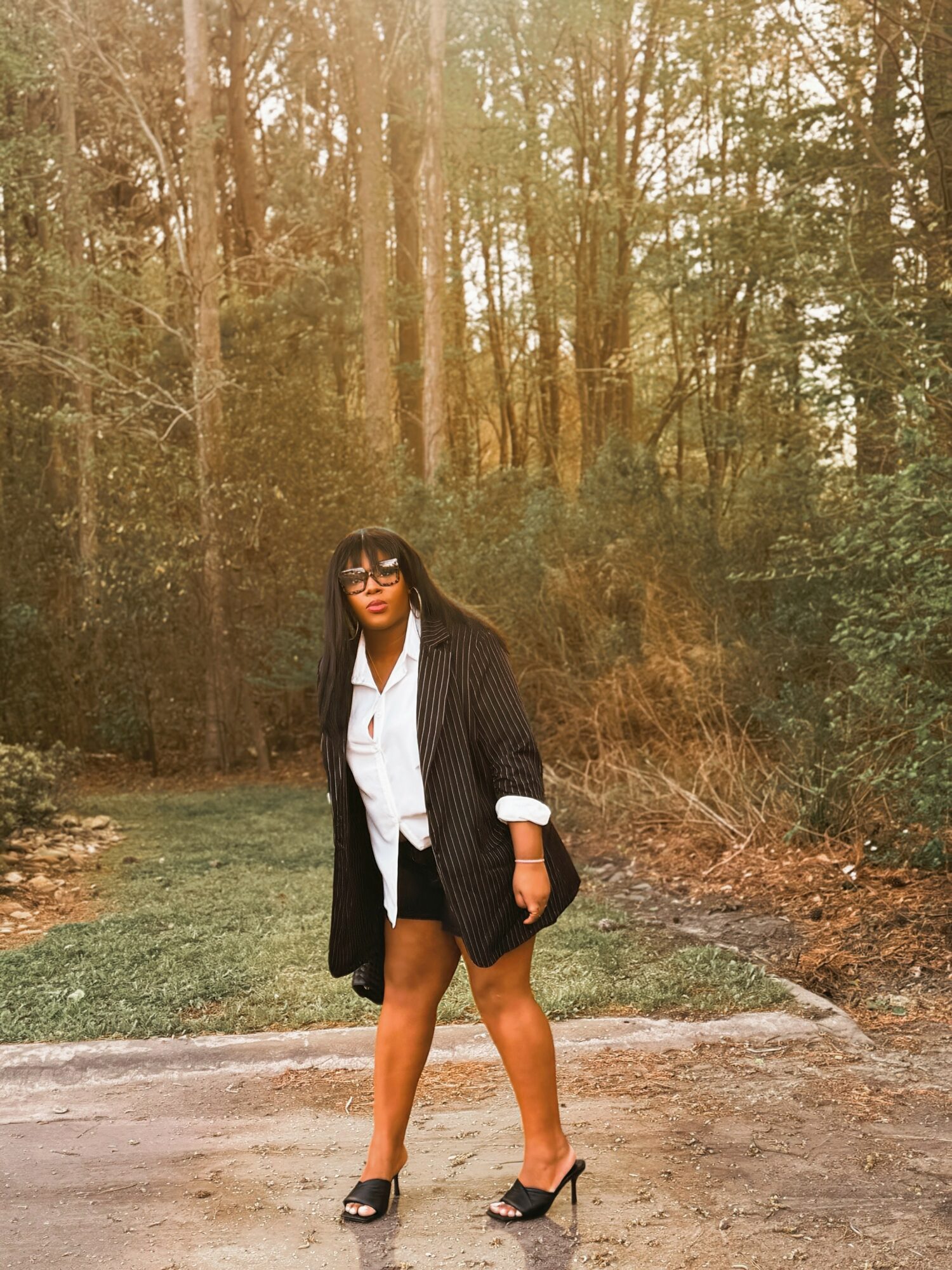 Woman standing outdoors in a forested area, wearing a black blazer, white shirt, shorts, and high heels.