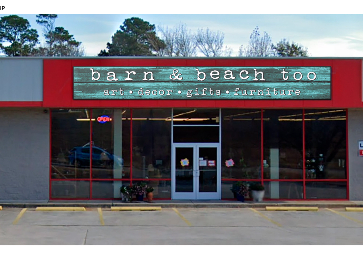 Storefront with a red frame, glass doors, and windows, displaying a sign for barn & beach too art & design gift boutique, with potted plants outside.
