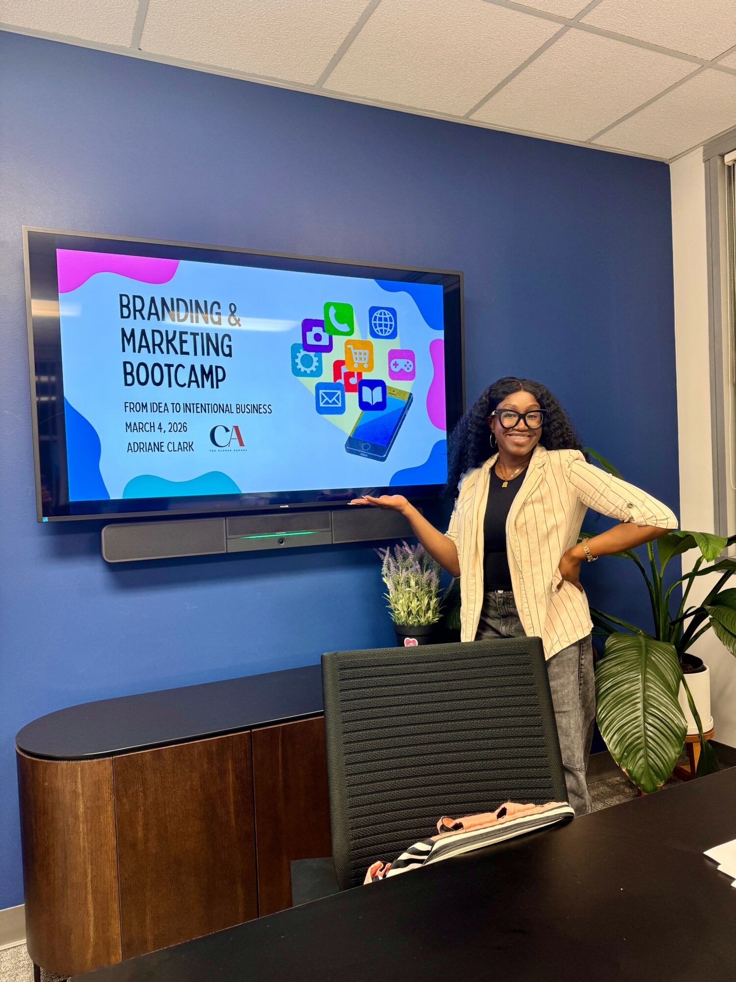 Woman standing next to a large screen displaying a presentation titled 'Branding & Marketing Bootcamp' in a conference room with plants and a table.