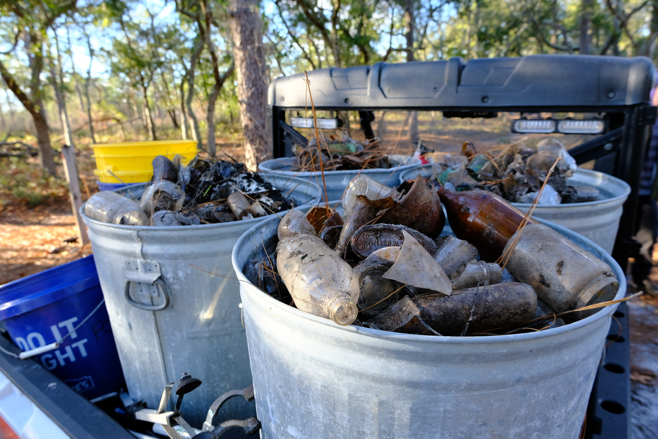 Two metal buckets filled with broken glass bottles outdoors, with trees and a vehicle in the background.