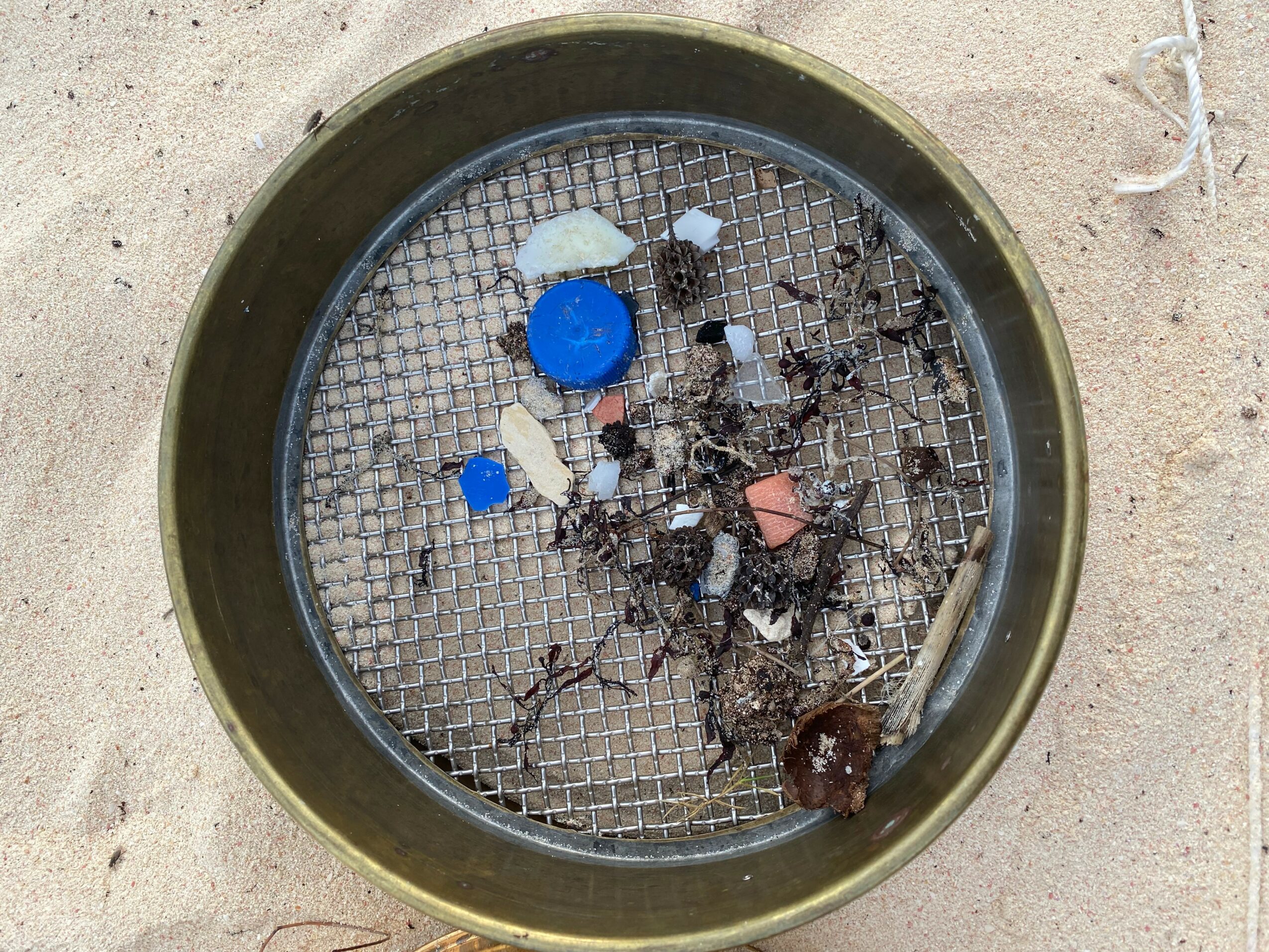 Round metal container with a mesh bottom, containing trash and debris, on a sandy surface.