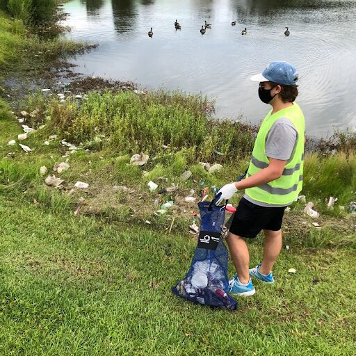 Person wearing a mask, hat, and reflective vest collecting trash near a lake with ducks swimming, holding a trash bag.