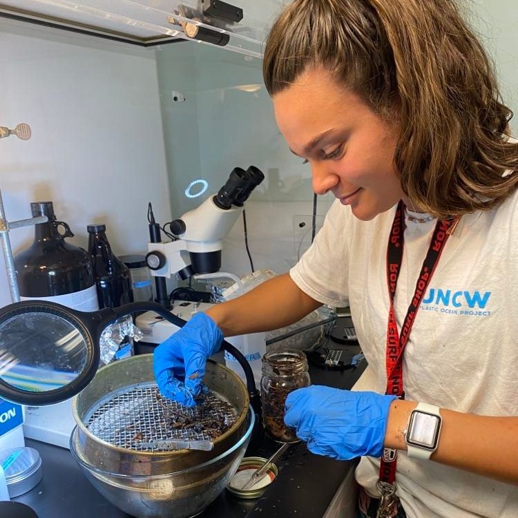 Woman in a lab working with a sieve and scientific equipment, wearing blue gloves and a white shirt with a logo.