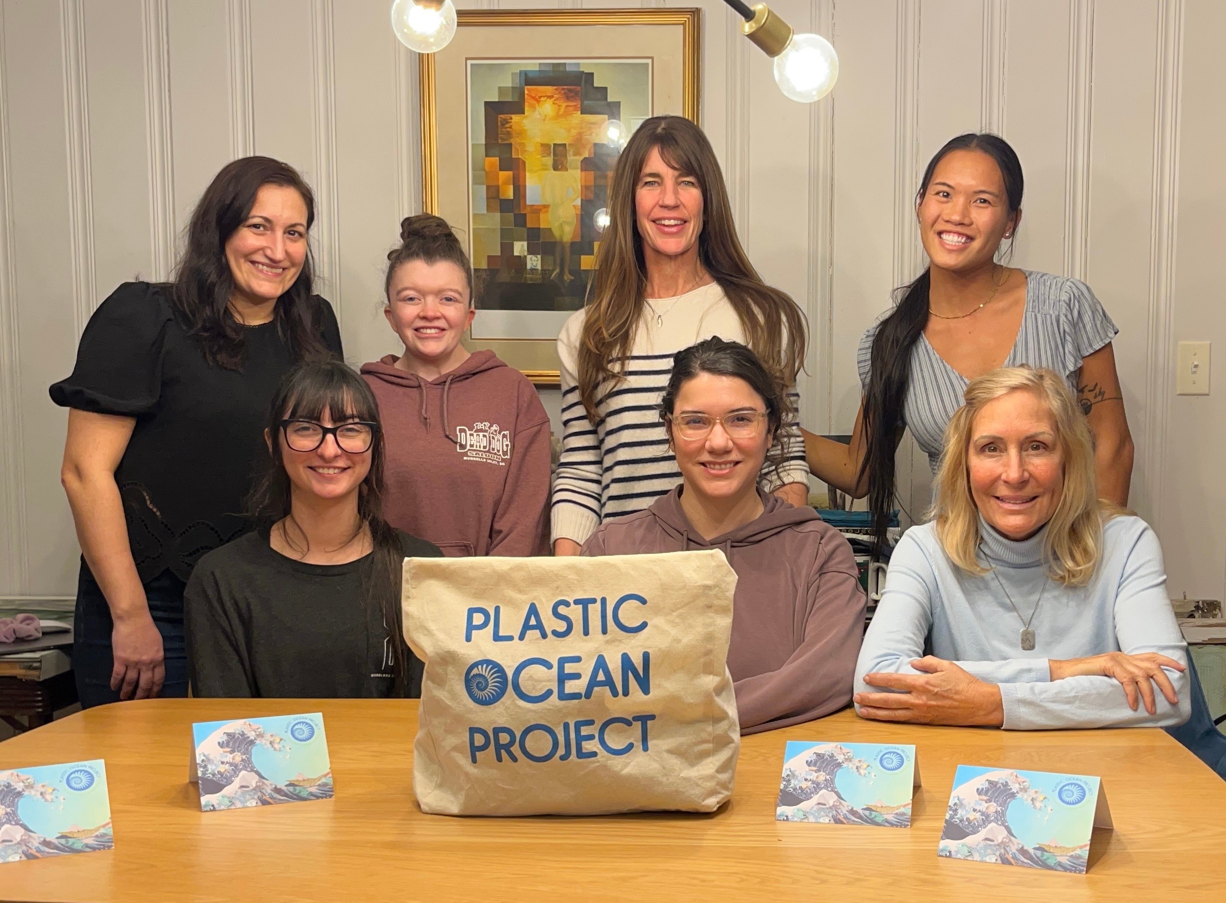 Seven people, including children and adults, sit and stand around a table with a sign that reads 'Plastic Ocean Project.'