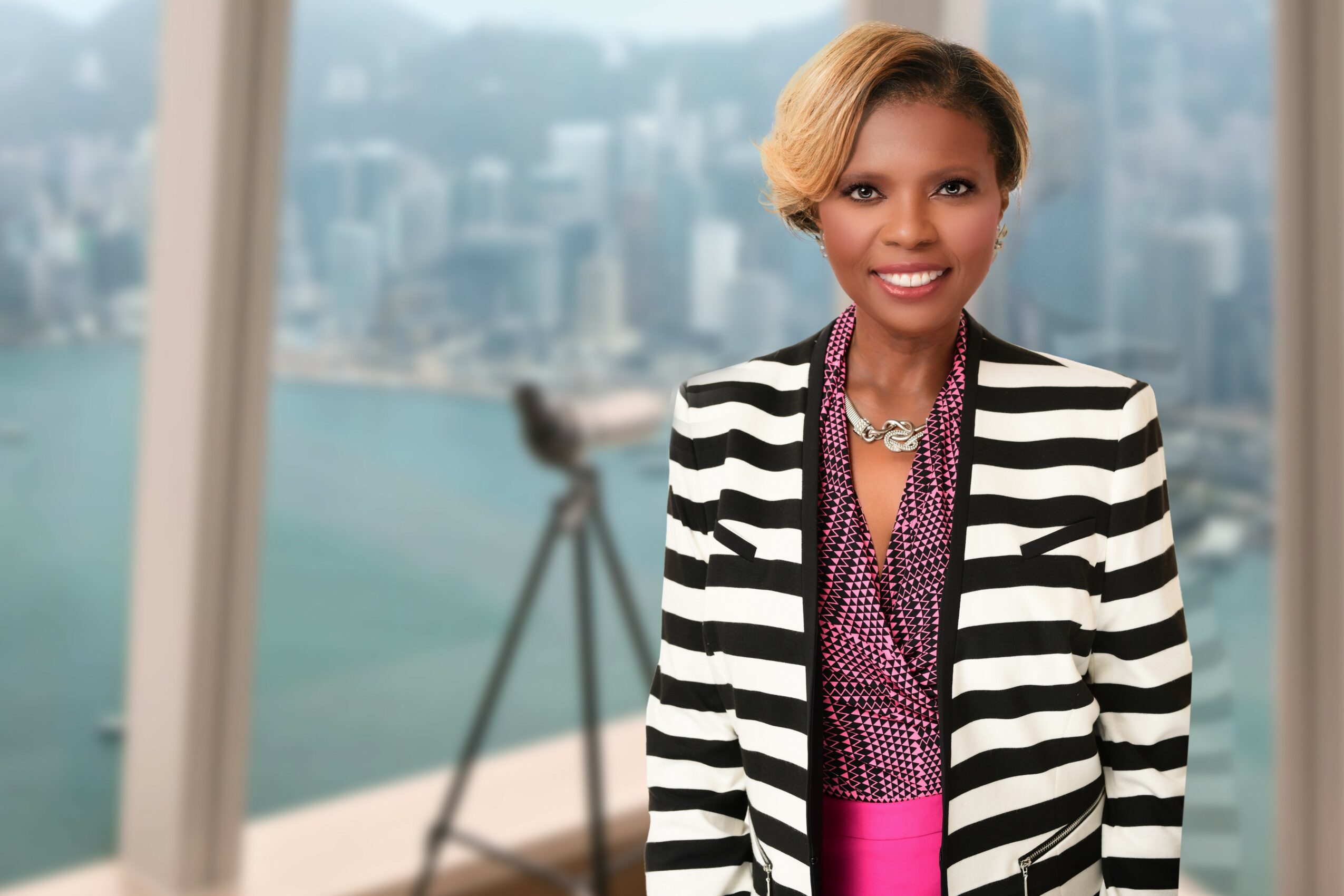 Smiling woman in a striped blazer and purple blouse stands in front of a cityscape with water and buildings in the background.