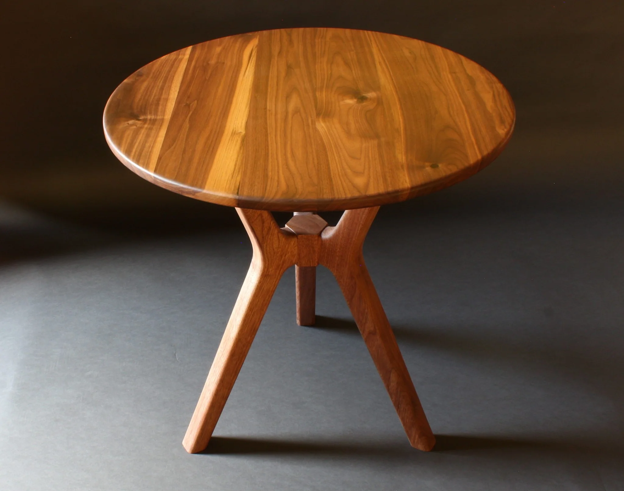 Round wooden table with three angled legs on a dark background.