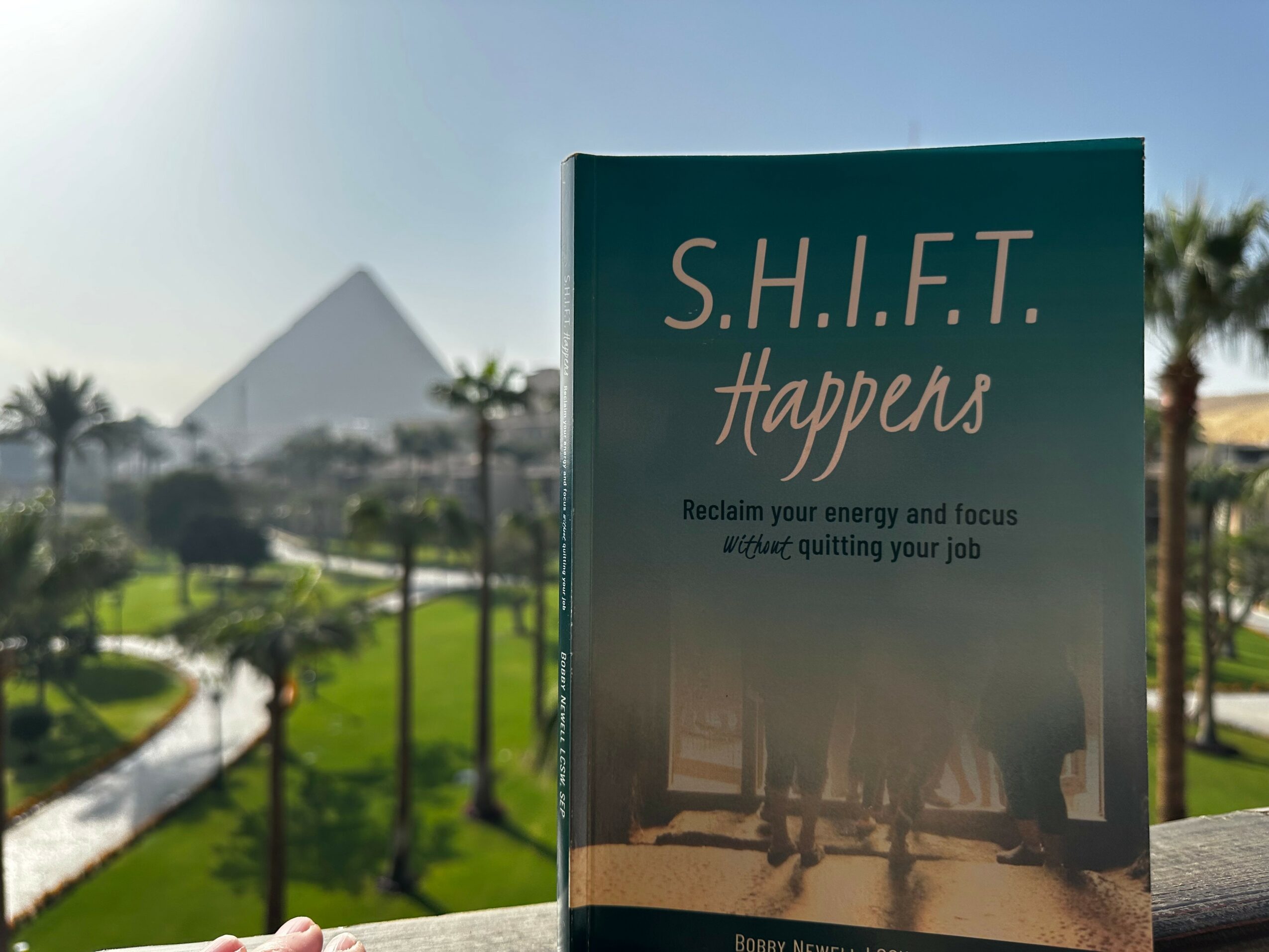 Book titled 'S.H.I.F.T. Happens' held outdoors with a park and pyramid in the background.