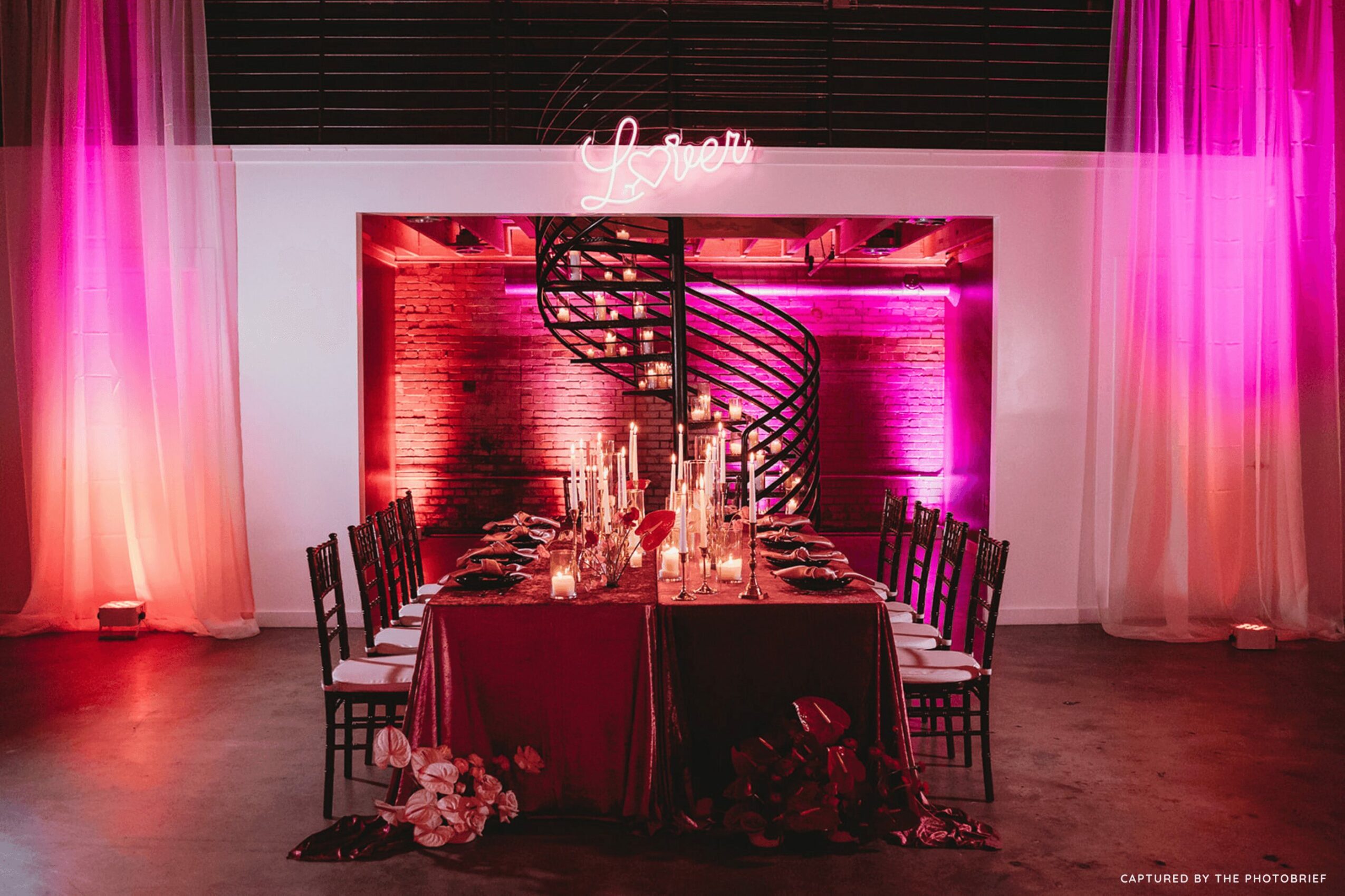 Elegant dining table set with candles and flowers, pink lighting, spiral staircase in background, pink curtains, and neon sign above.