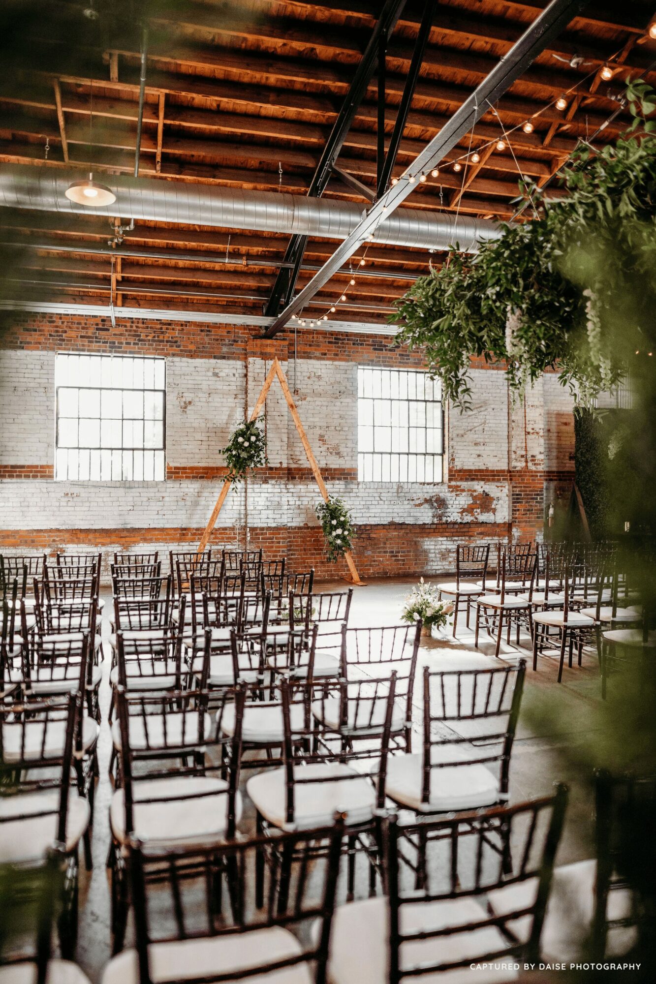 Indoor event space with rows of chairs, large windows, brick walls, and hanging greenery, ready for a gathering.