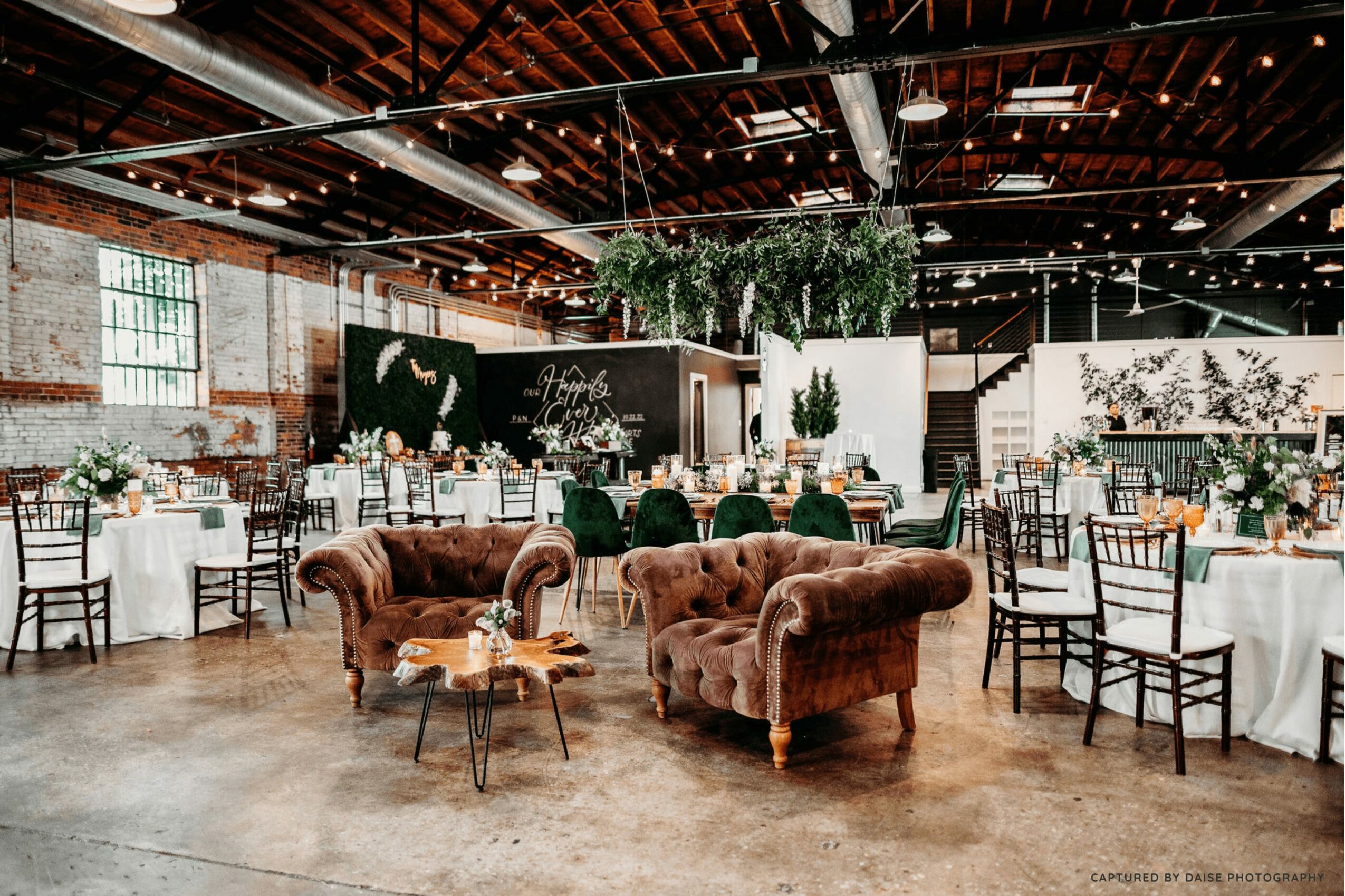 Elegant event space with round tables, chairs, and plush armchairs, decorated with string lights and greenery, in a brick-walled venue.