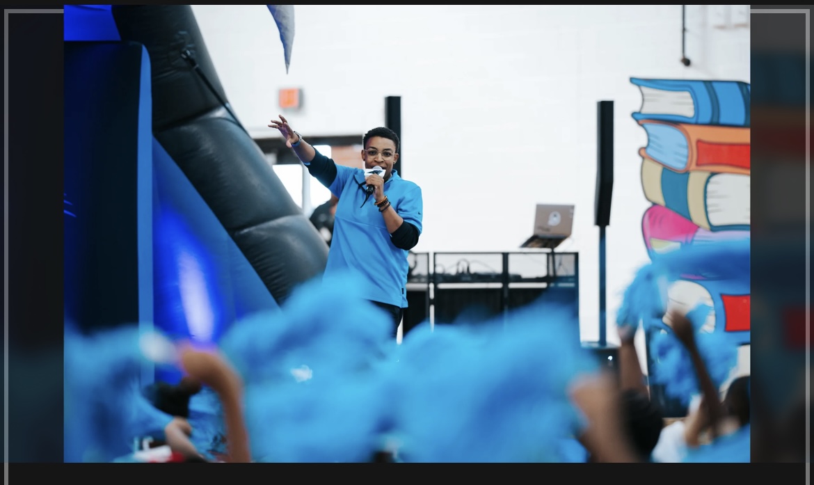 A person in a blue shirt speaking into a microphone on stage, with a colorful book sculpture in the background.