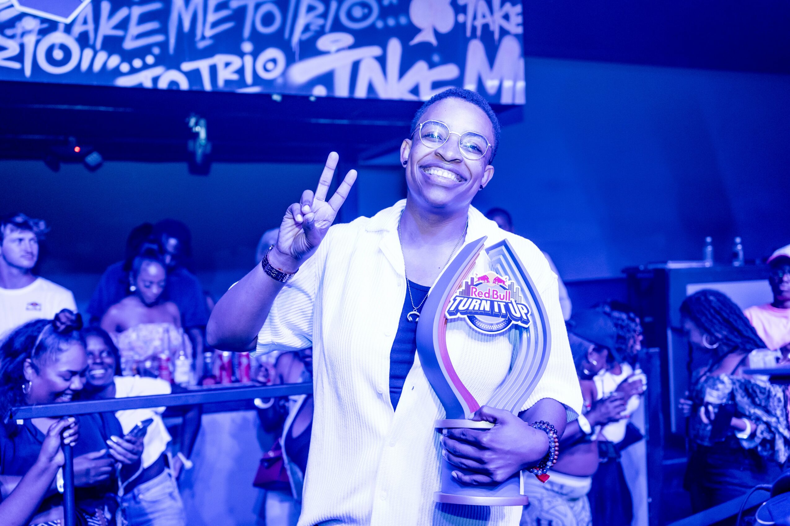 Smiling person holding a trophy and making a peace sign, surrounded by people in a lively indoor event.
