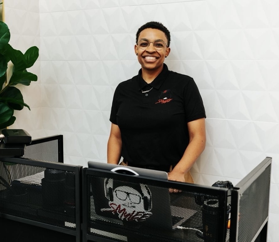 Smiling woman standing behind a reception desk in an office with a white textured wall and a green plant.