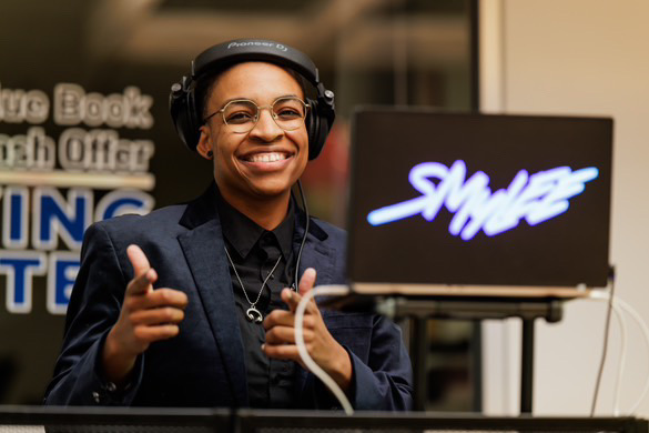 Smiling woman wearing headphones and glasses, giving a thumbs-up, in front of a laptop with a stylized logo.