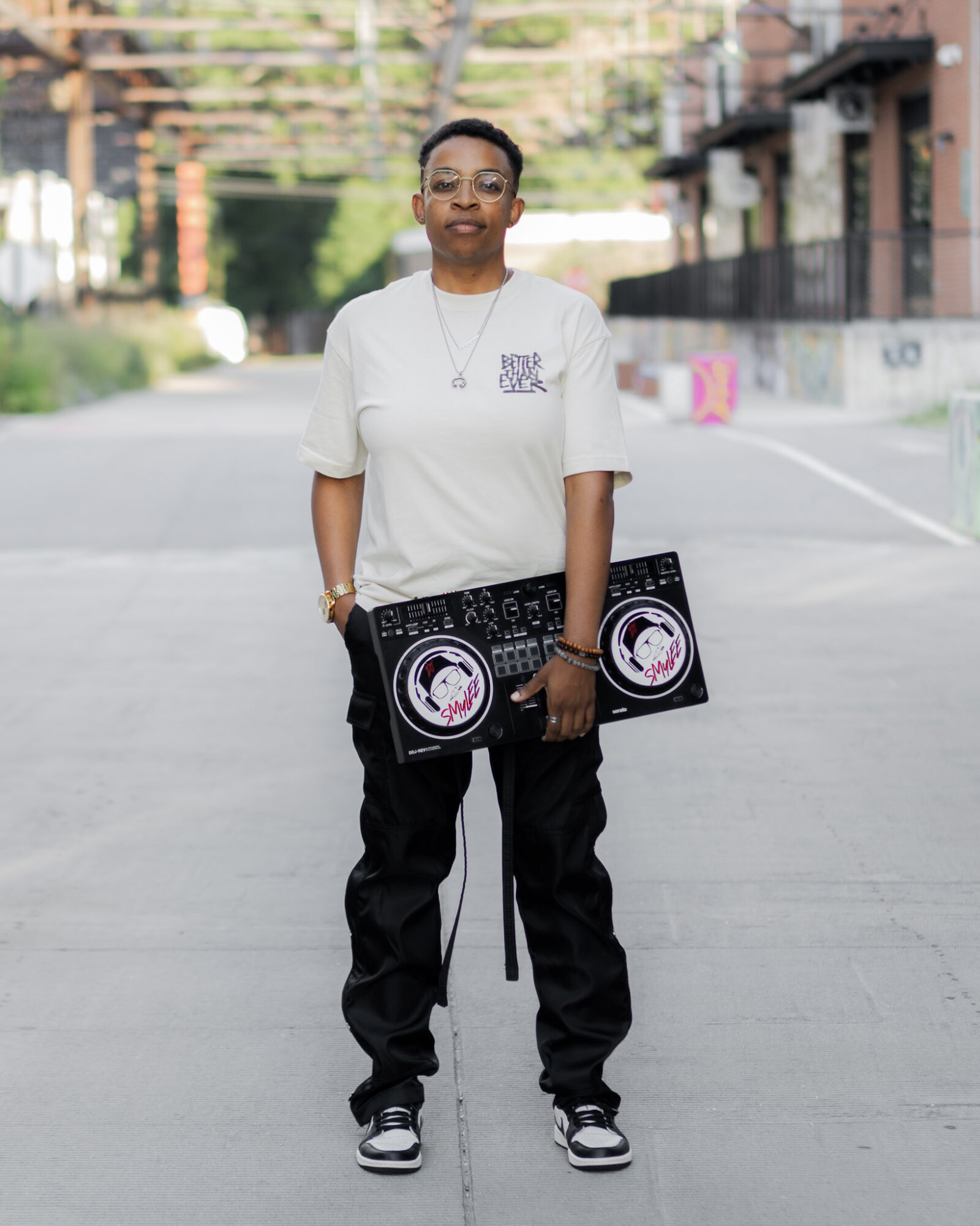 Person standing on street holding two large black speakers, wearing a white t-shirt, black pants, and sneakers.