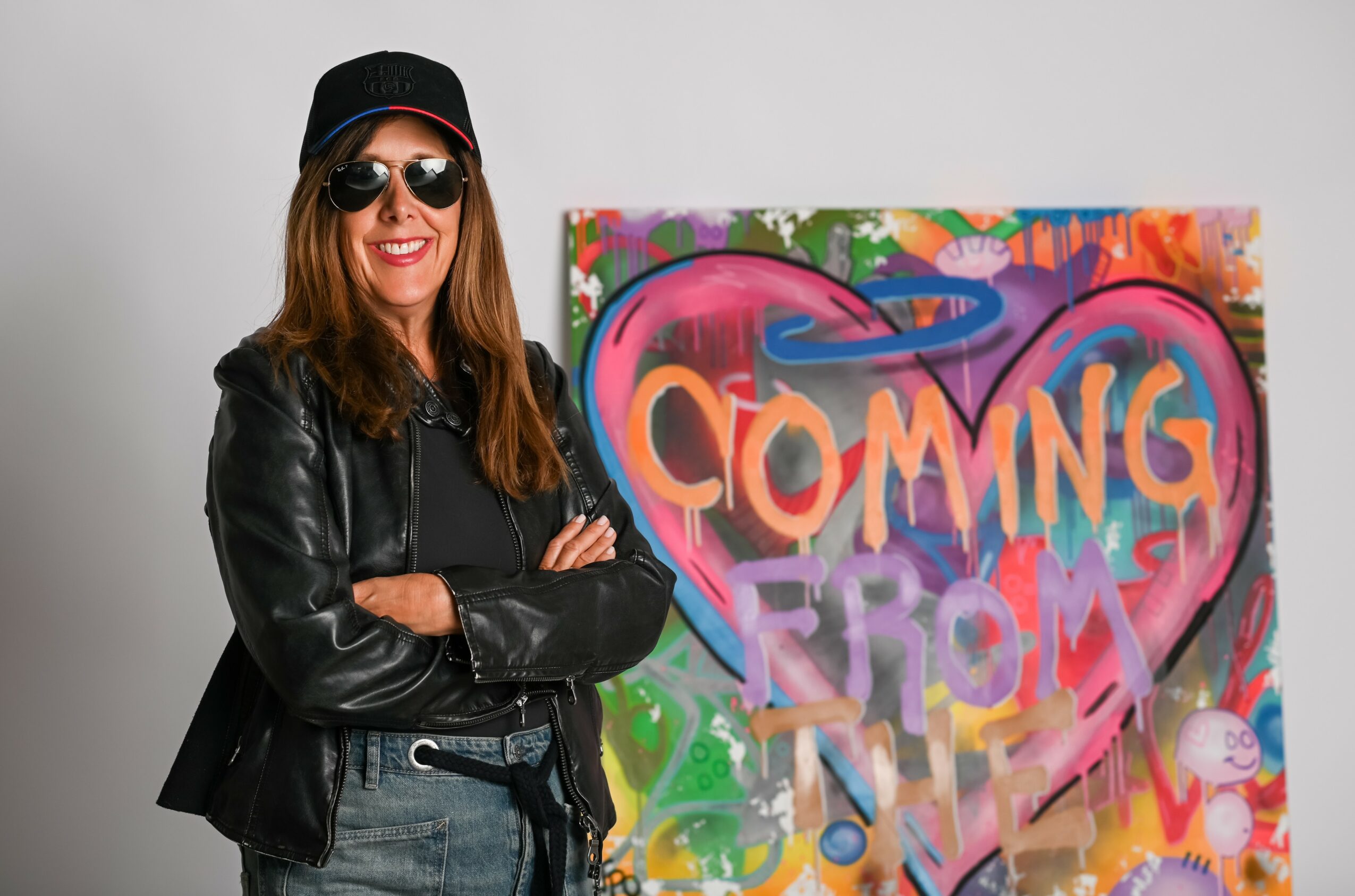 Woman with sunglasses and a leather jacket standing next to colorful graffiti art with the words 'COMING FROM' inside a heart shape.