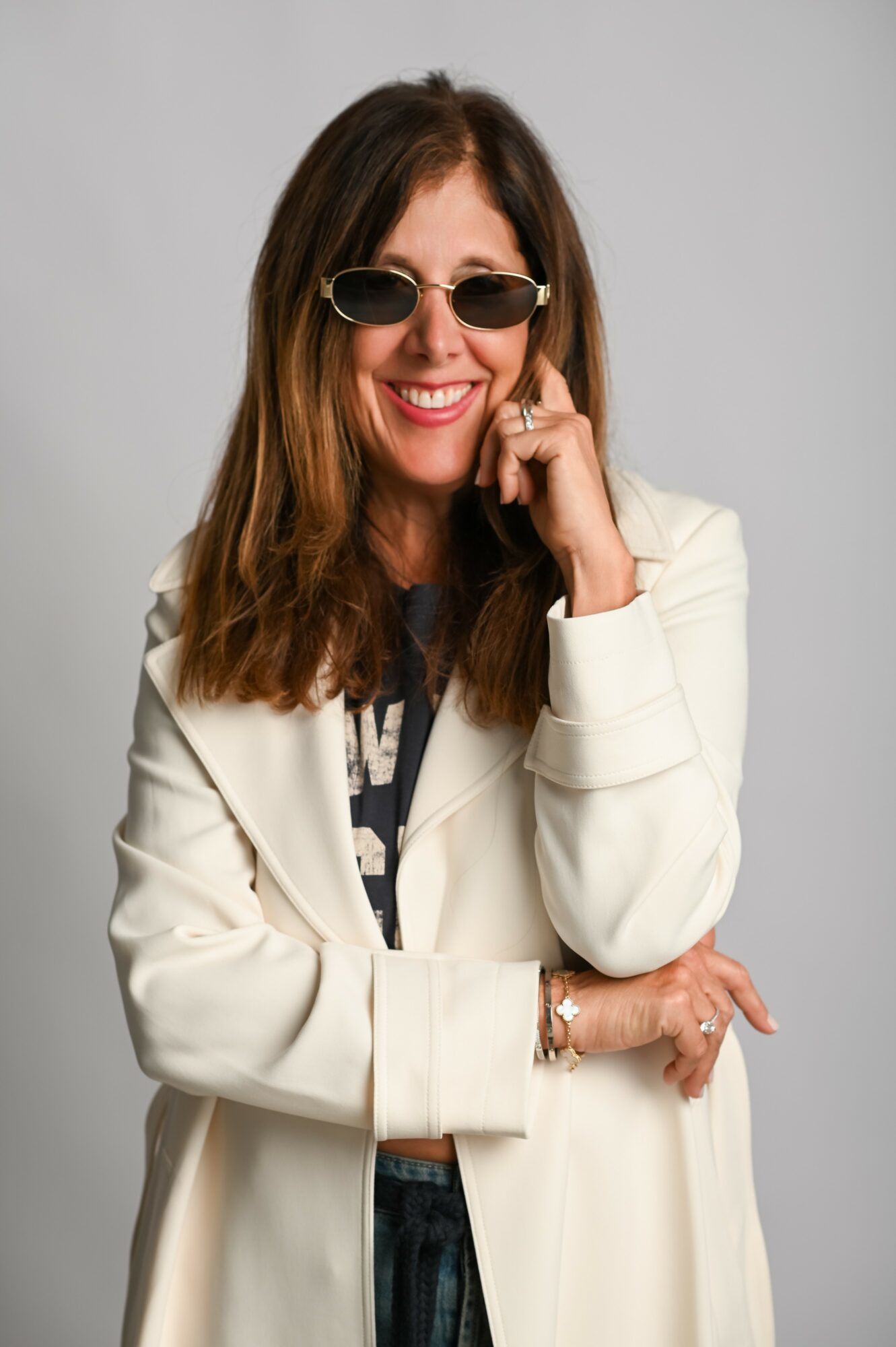 Woman with long brown hair wearing sunglasses, a white blazer, and smiling, with her hand near her face.