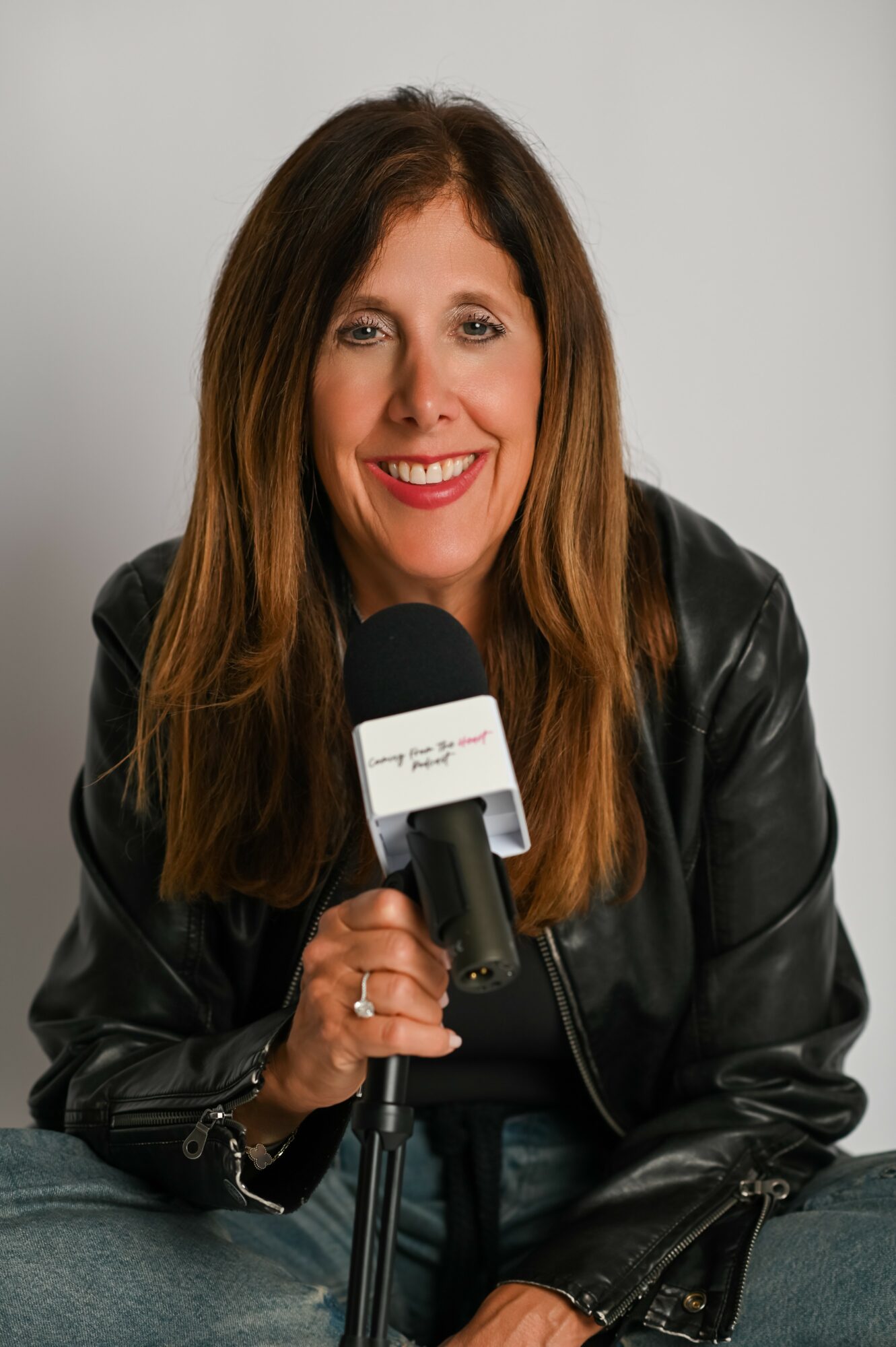 Woman with long brown hair smiling, holding a microphone, wearing a black leather jacket, sitting against a plain background.