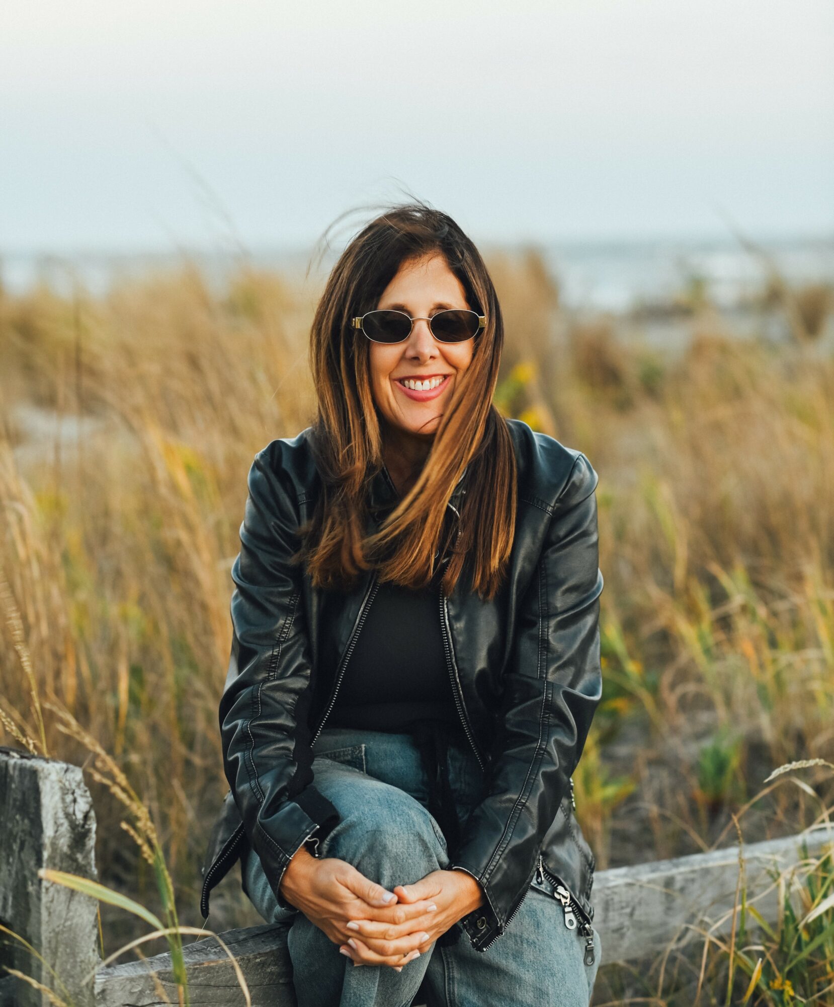 Woman with long hair wearing sunglasses and a black leather jacket, sitting outdoors in a grassy area.