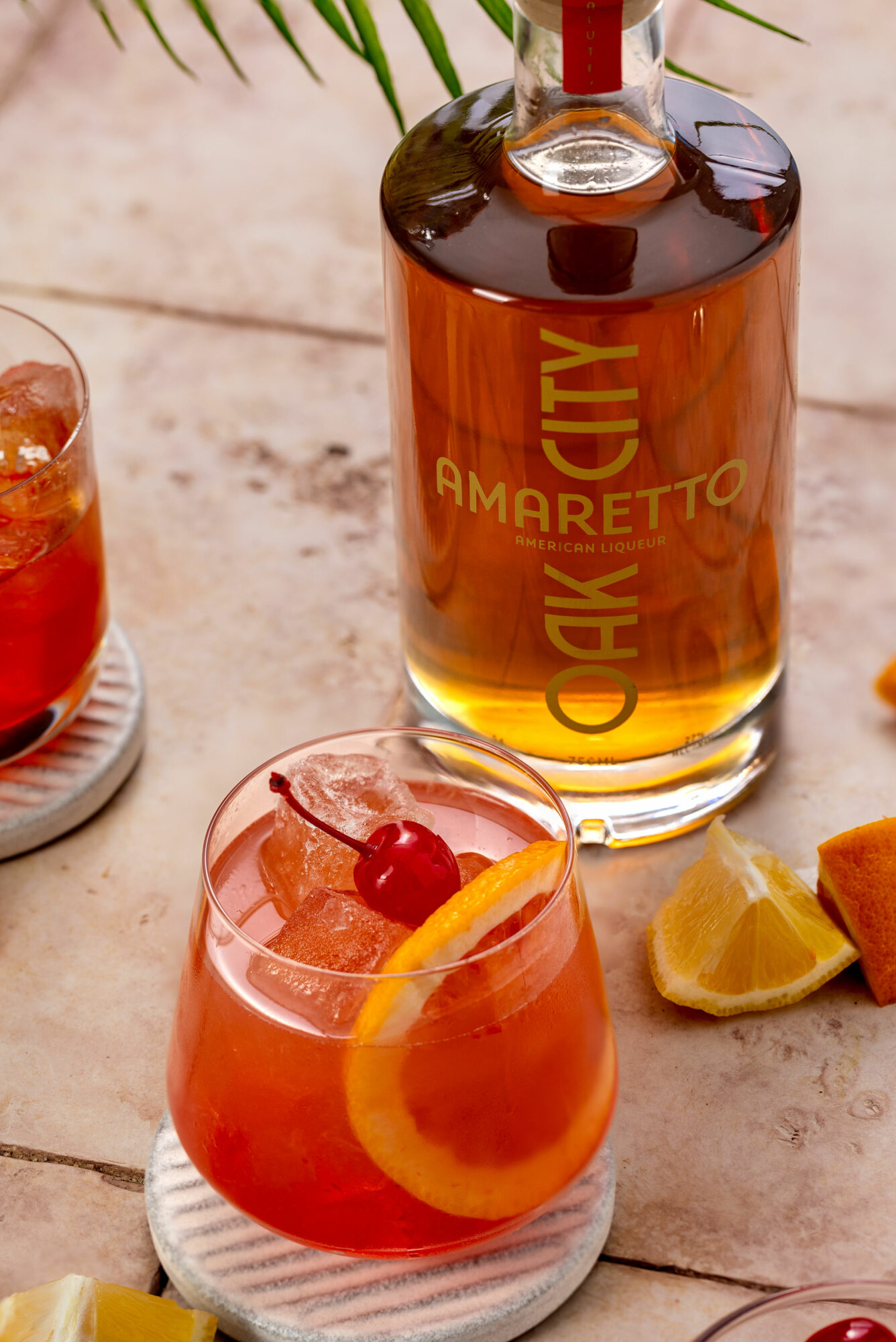 Bottle of Oak City Amaretto liqueur, a cocktail with ice, lemon, and cherry, and lemon wedges on a wooden surface.