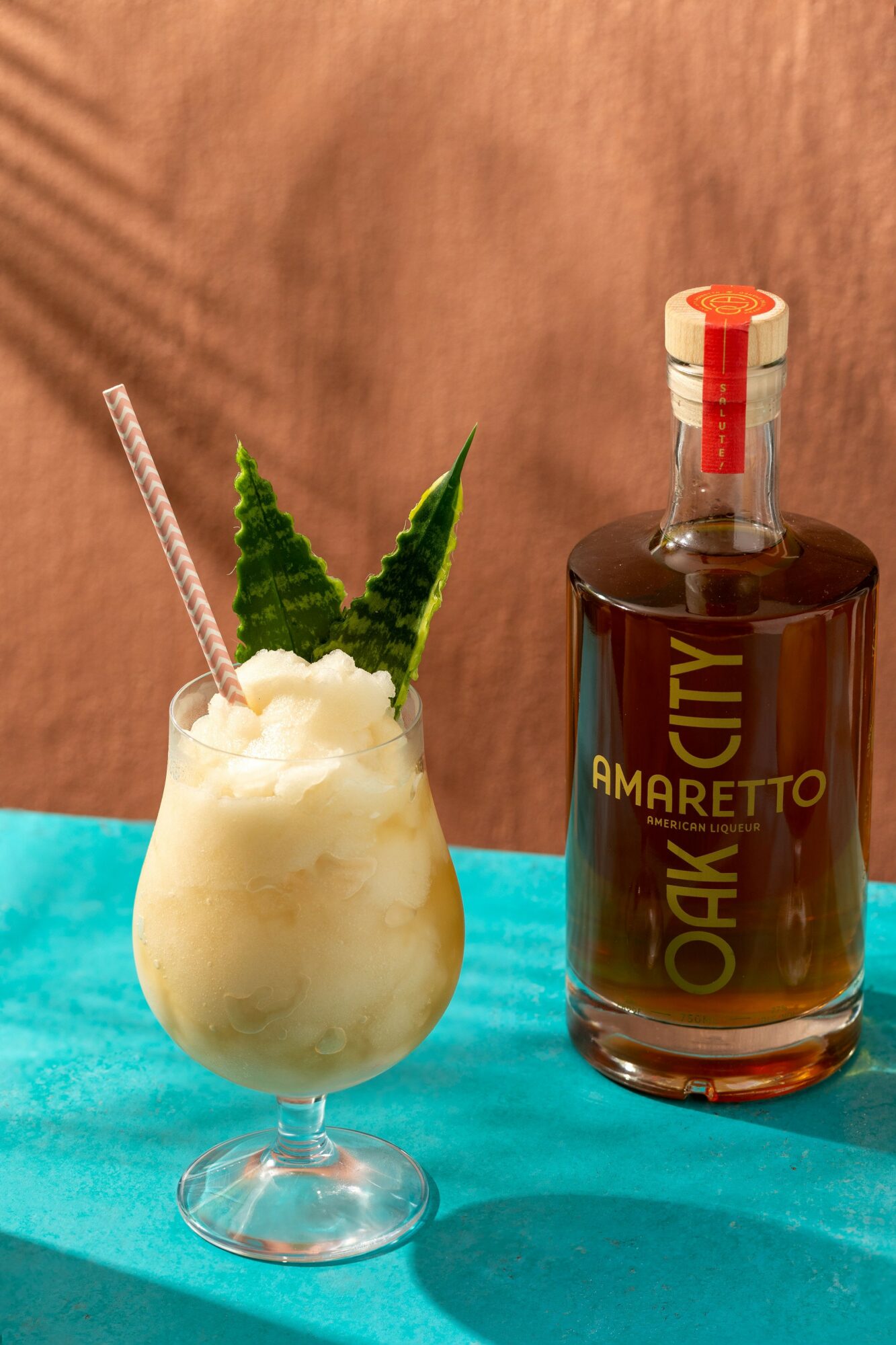 A glass with a creamy drink, a straw, and a green leaf, next to a bottle with a red cap on a turquoise surface.