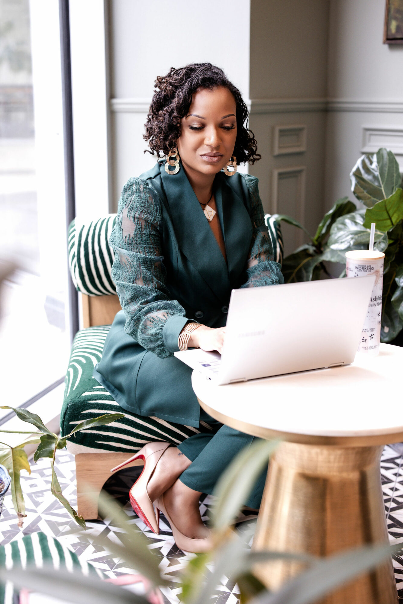 Woman with curly hair working on a laptop at a round table in a bright room with plants.