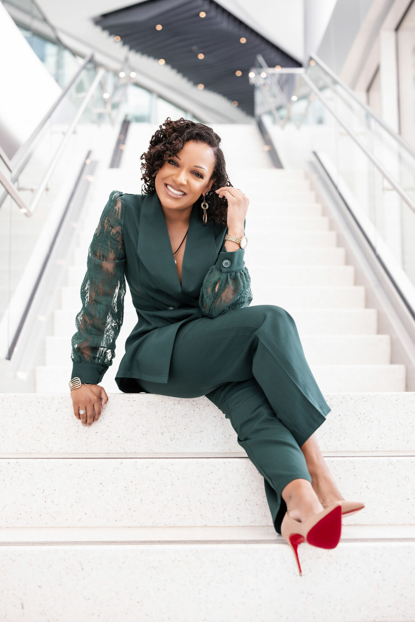 Woman sitting on stairs, smiling, wearing a green outfit and red high heels, in a modern building interior.