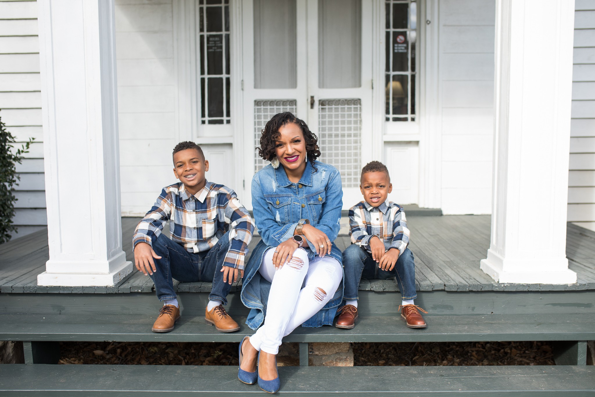 Woman sitting on porch with two children, all smiling, in front of a white house with columns.