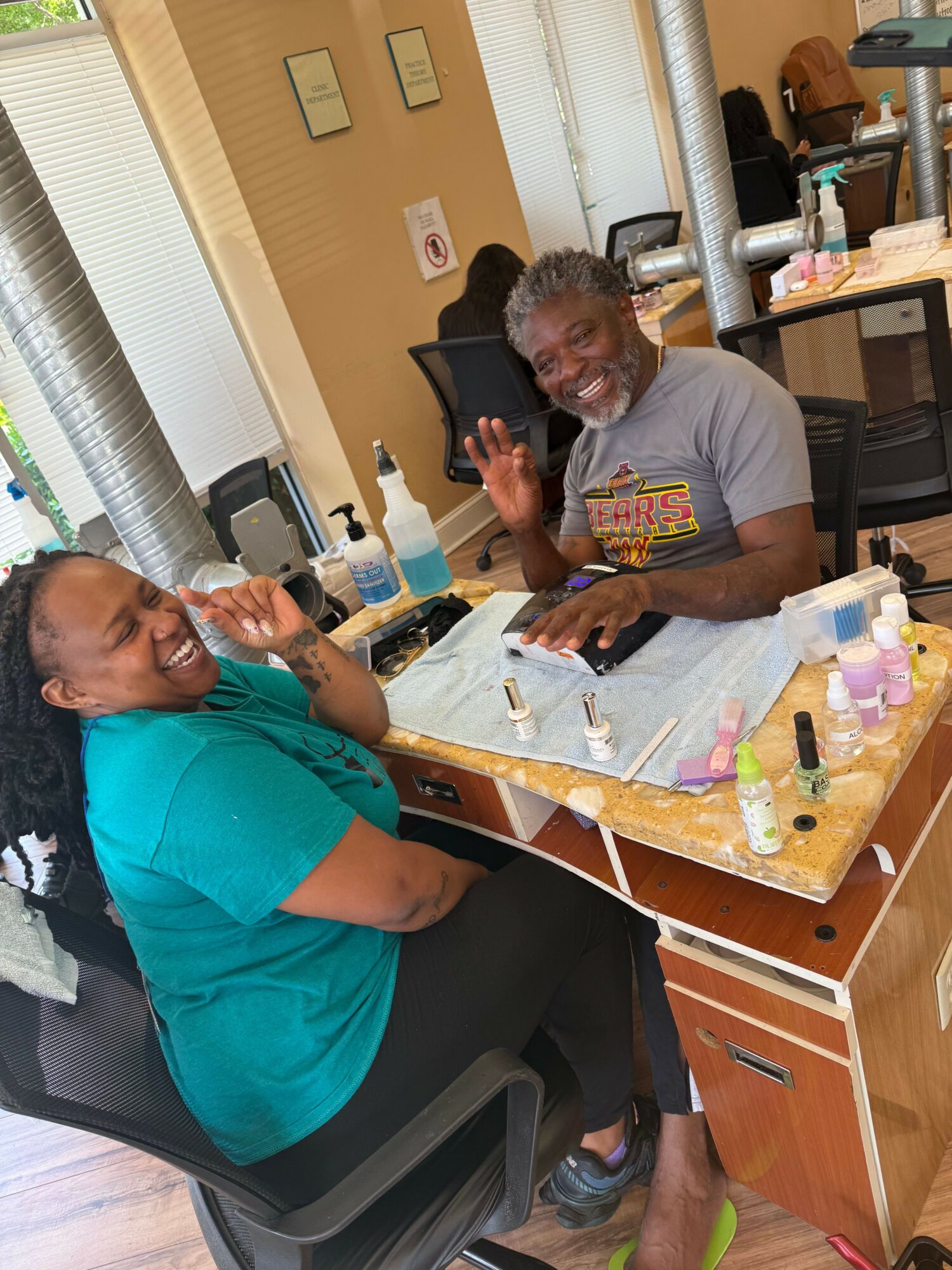 Two people sitting at a table with various bottles and items, smiling and looking at the camera.