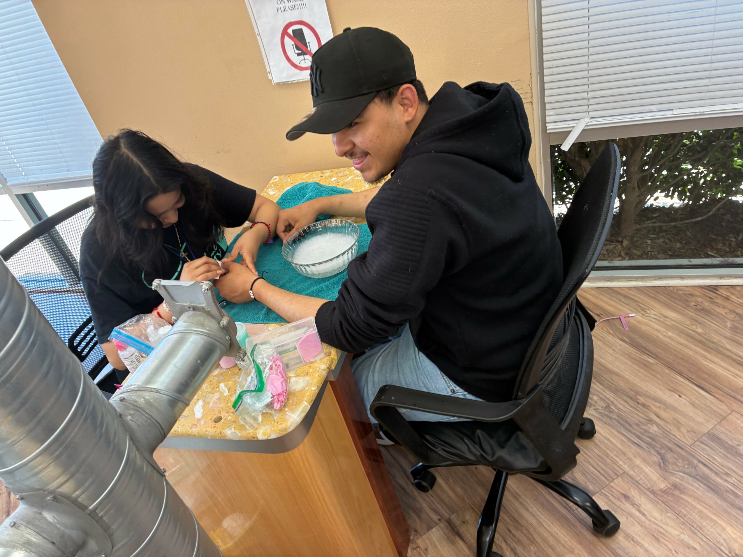 A person with long dark hair and a black shirt receives a tattoo from a person in a black hoodie, seated at a table. Sunlight streams through windows.