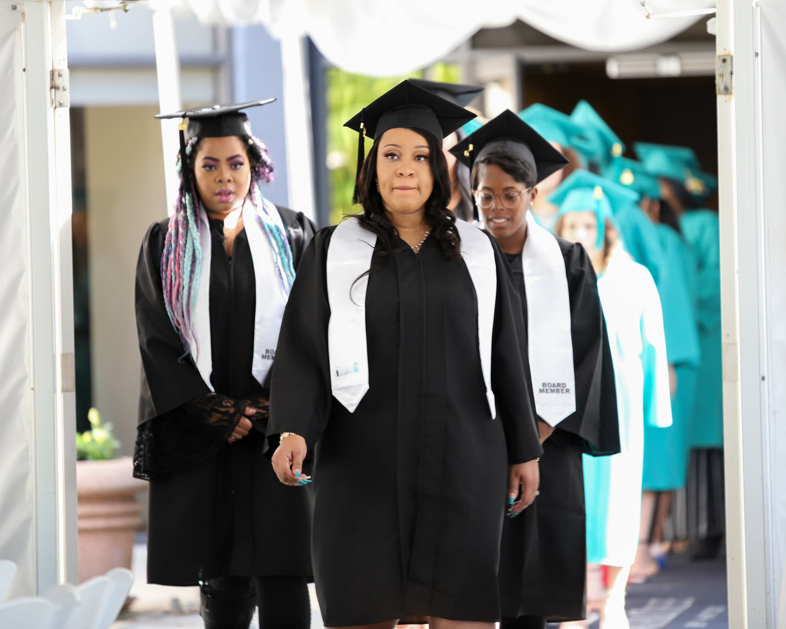 Group of graduates in black gowns and caps walking outdoors, some with white stoles, with a line of graduates in blue gowns behind them.