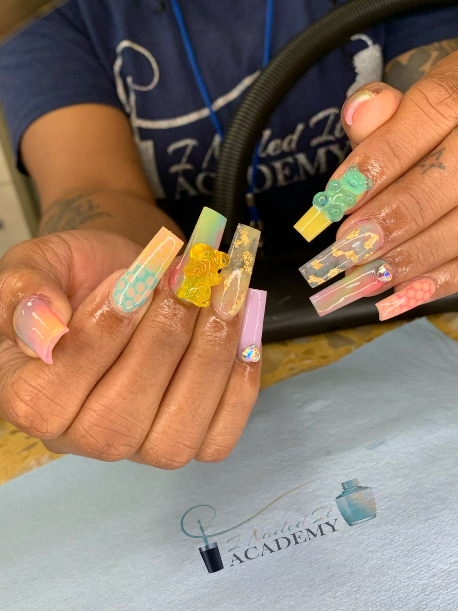 Close-up of hands with colorful, decorated acrylic nails, holding a nail art tool, with a logo on paper below.