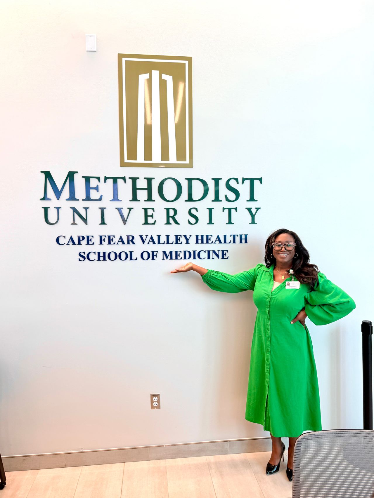 Woman in green gown standing next to Methodist University sign at Cape Fear Valley Health School of Medicine.