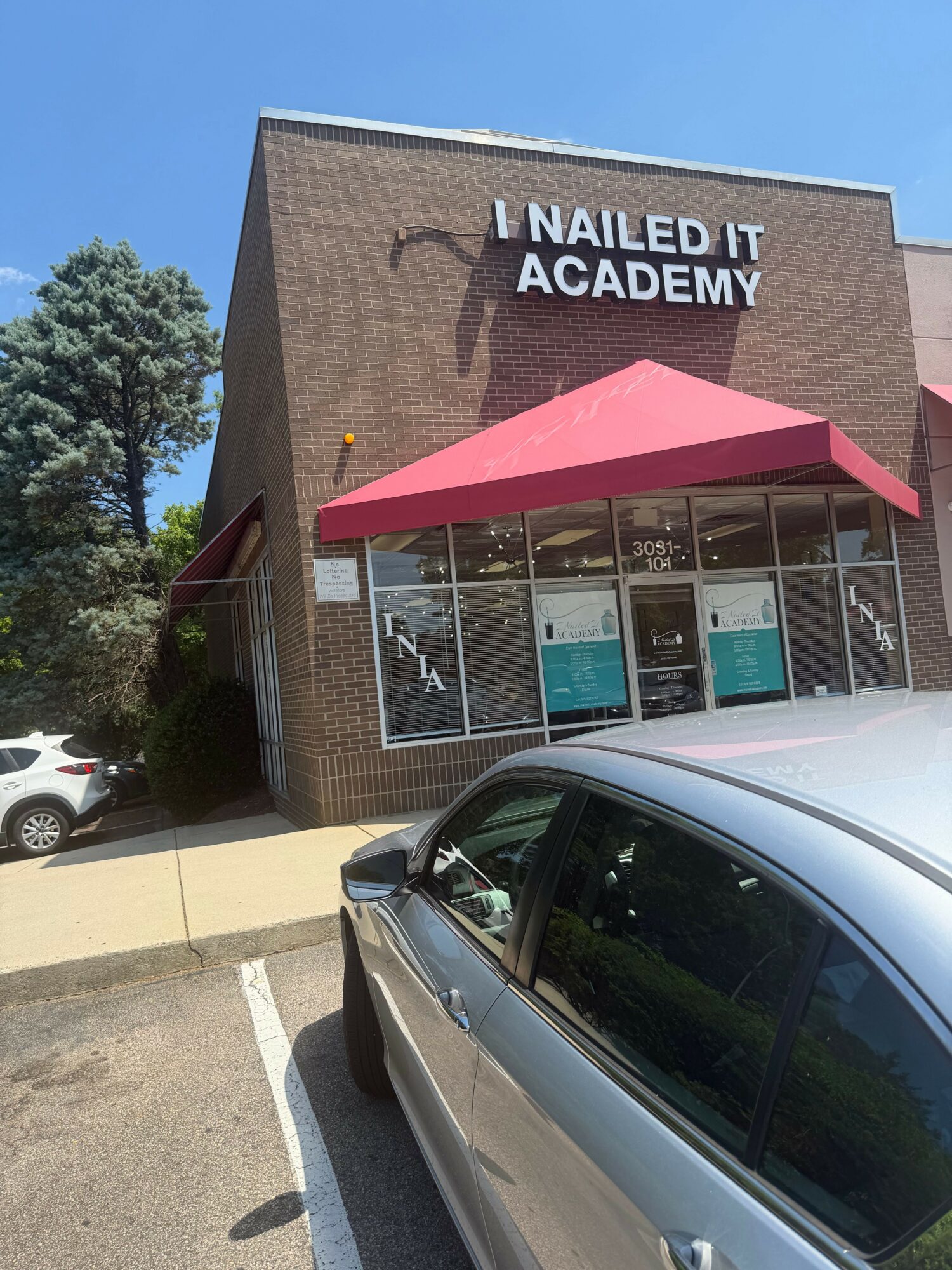 Nailed It Academy building with glass windows and a red awning, parked cars in front, blue sky above.