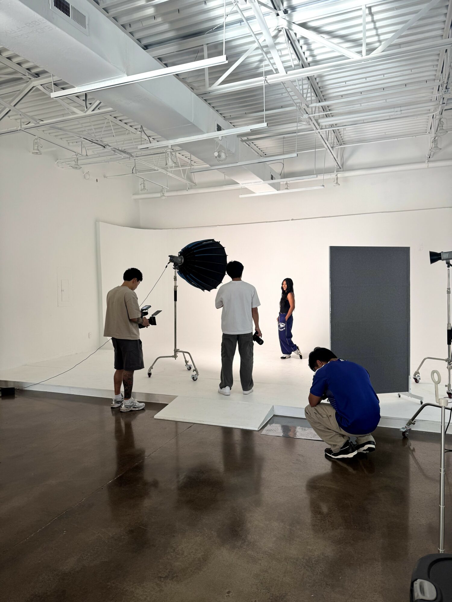 Four people in a photography studio with a white backdrop, lighting equipment, and a gray panel, some adjusting equipment and posing.