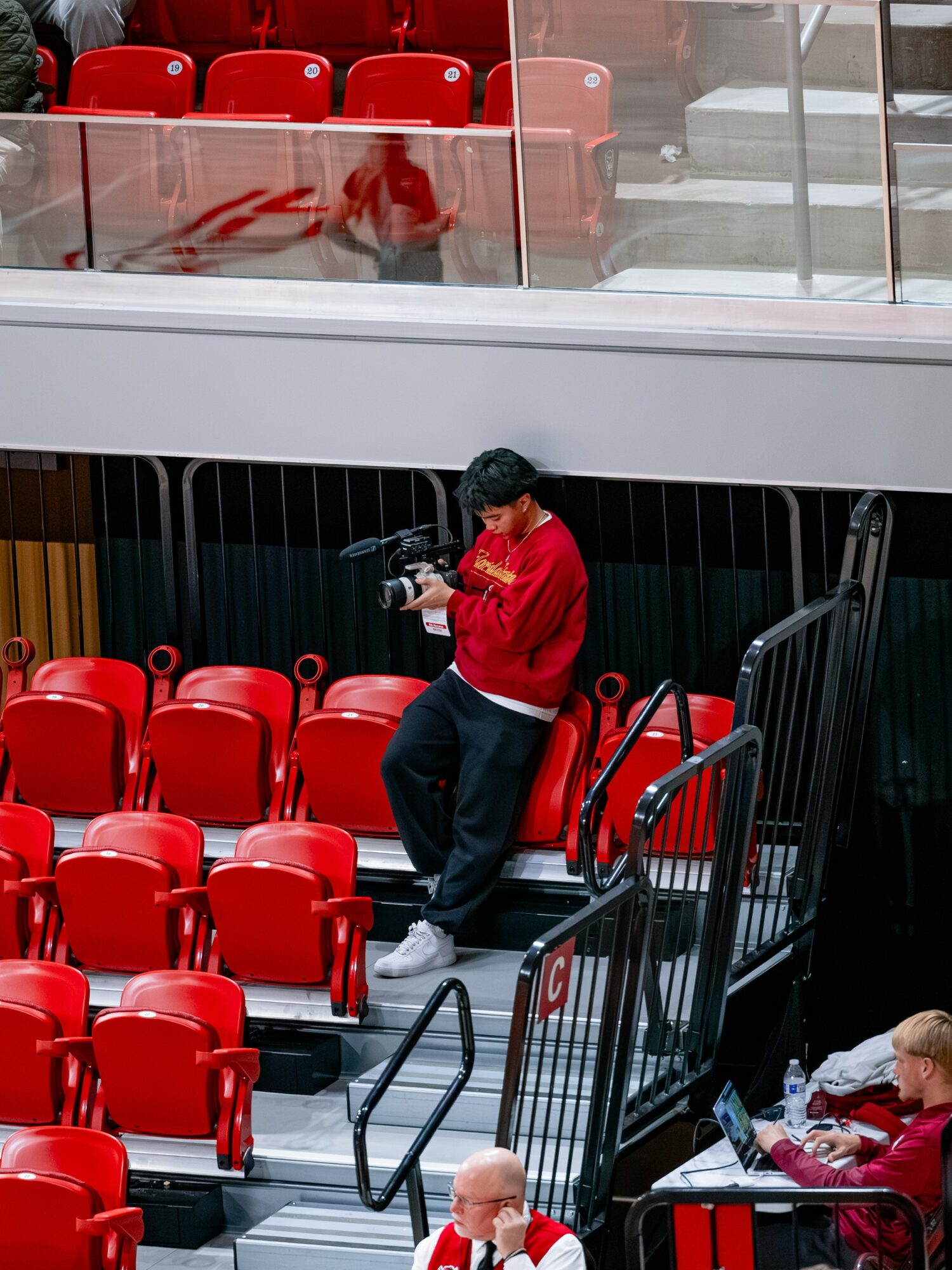 Person in red jacket standing on stairs, looking at a device, surrounded by red seats in an indoor setting.