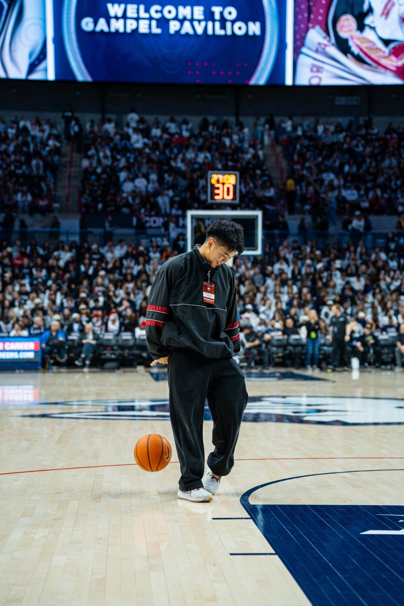 Person standing on basketball court, holding a basketball, with a crowd and scoreboard in background.