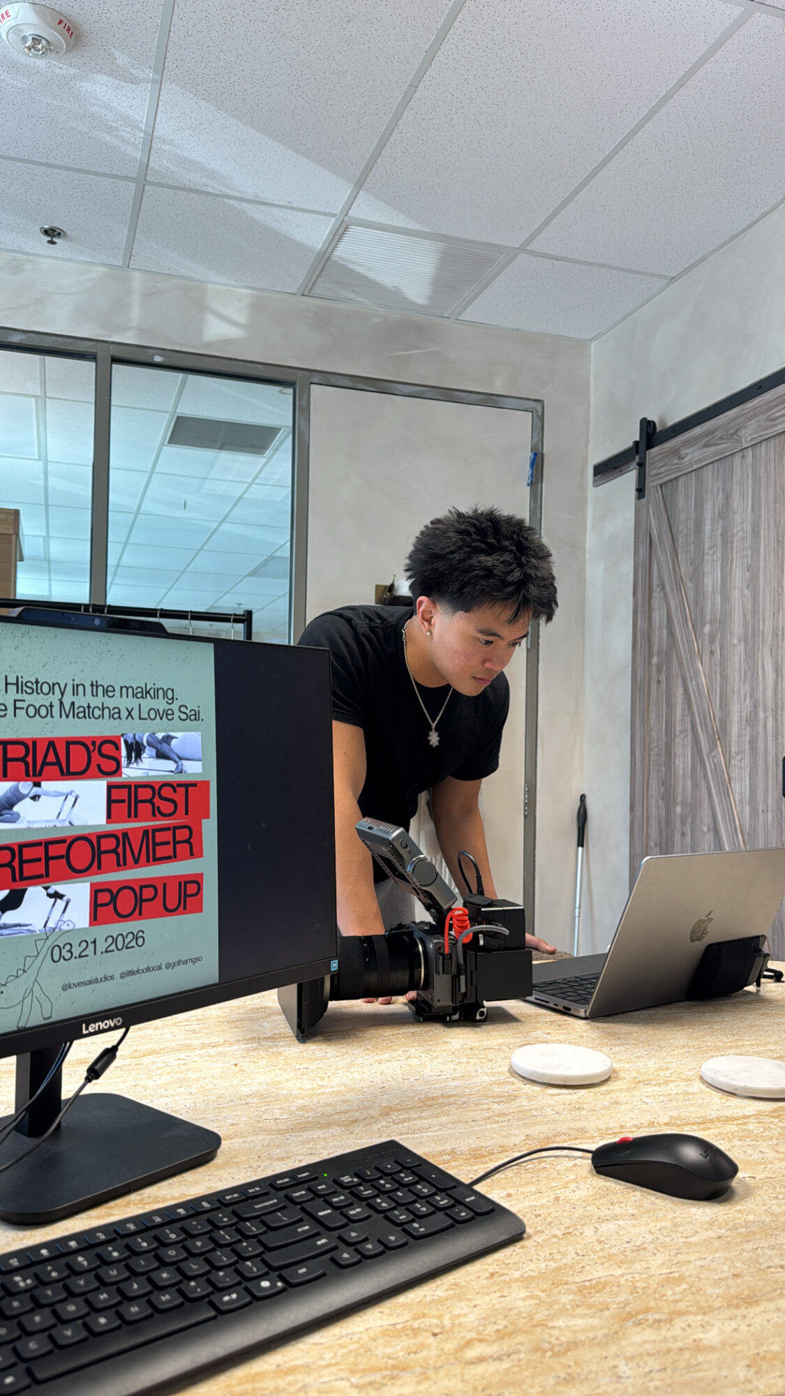 Person with curly hair working with camera equipment at a table, computer monitor and laptop nearby, in a modern office.