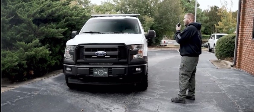 Man taking photo of a white pickup truck parked on a paved area with trees and bushes nearby.