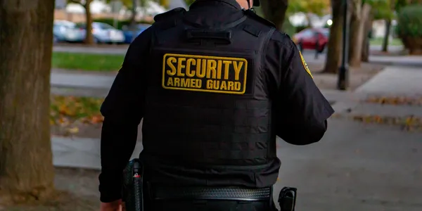 Security guard wearing a black vest with yellow text, standing outdoors on a sidewalk, trees and parked cars in background.