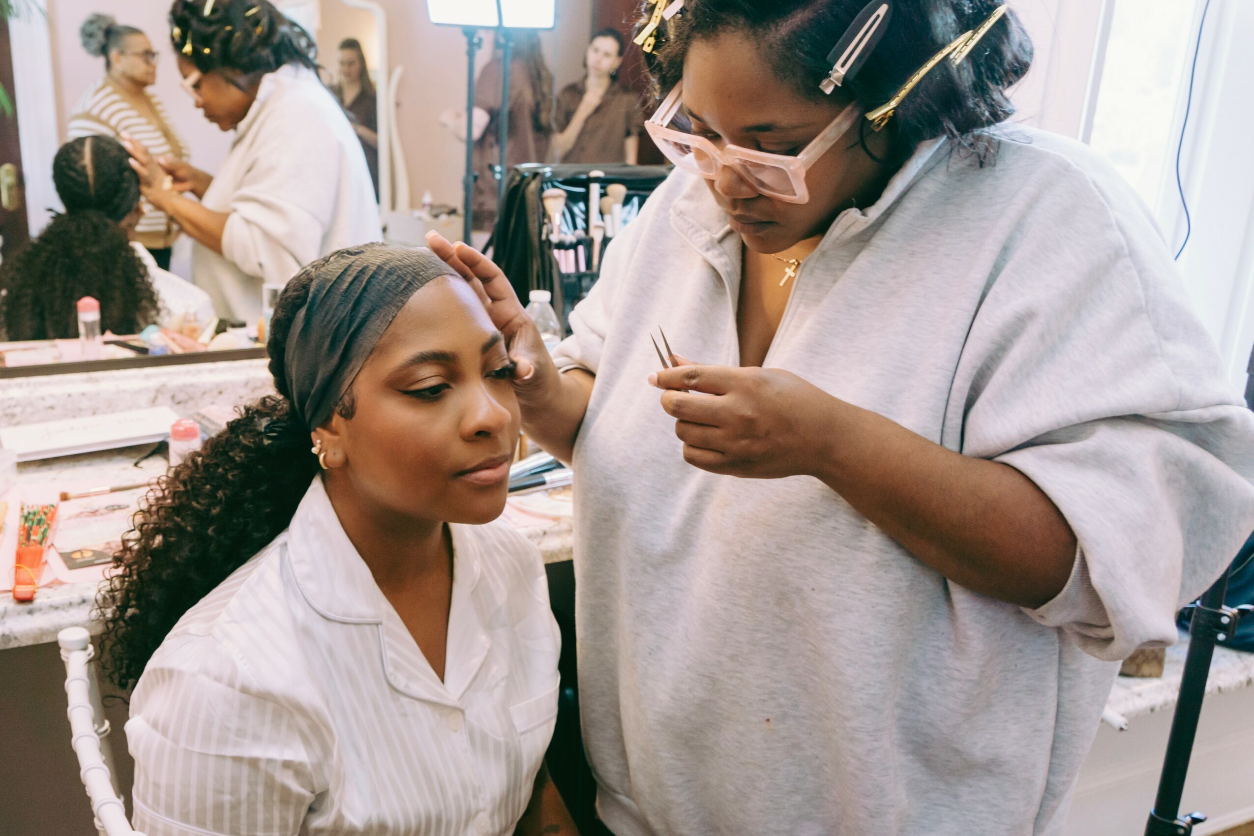 Makeup artist applying makeup to a woman in a beauty salon, with other clients and staff in the background.