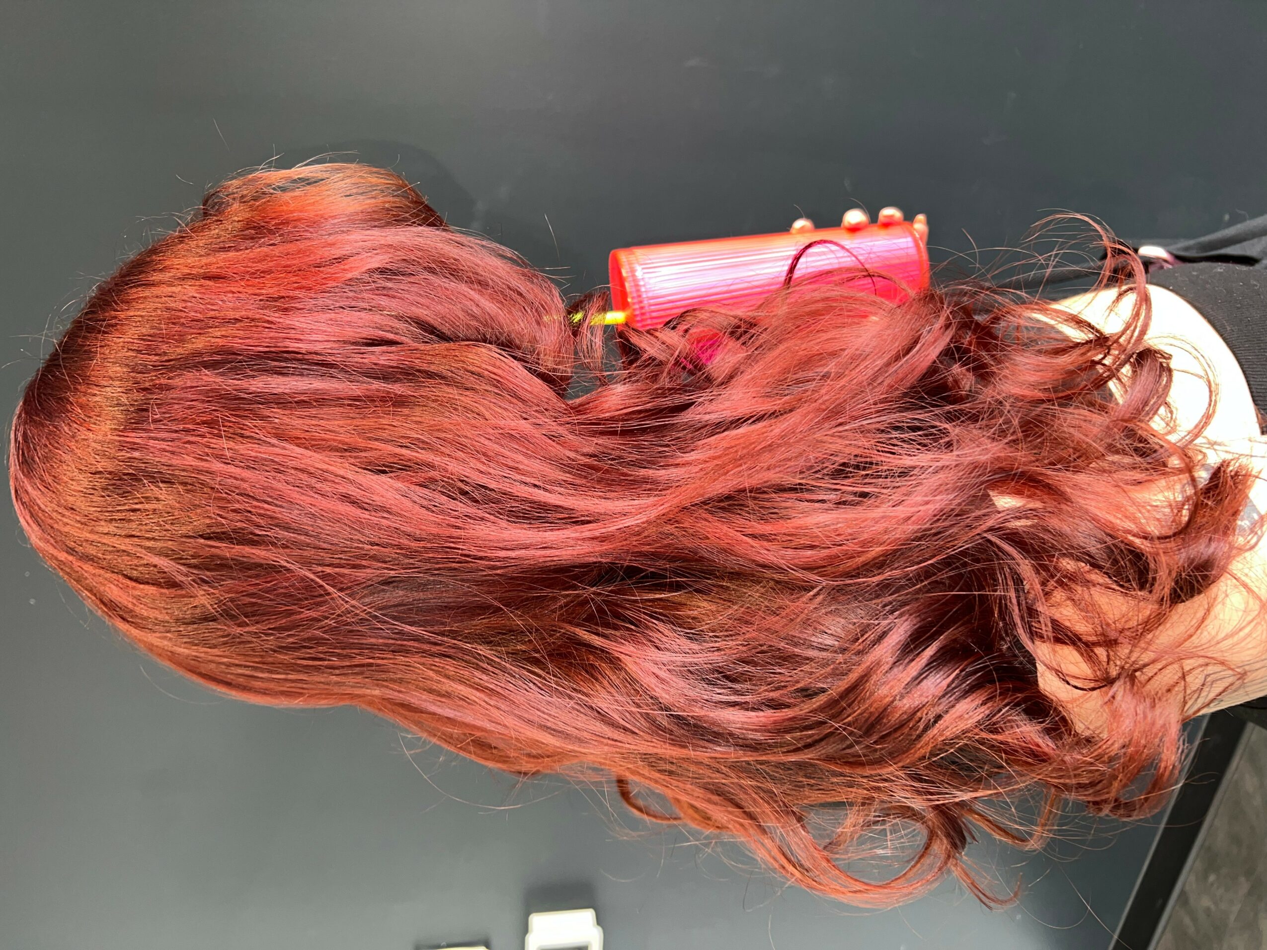 Person with long, wavy, reddish hair lying on a surface, with a pink hair roller nearby.