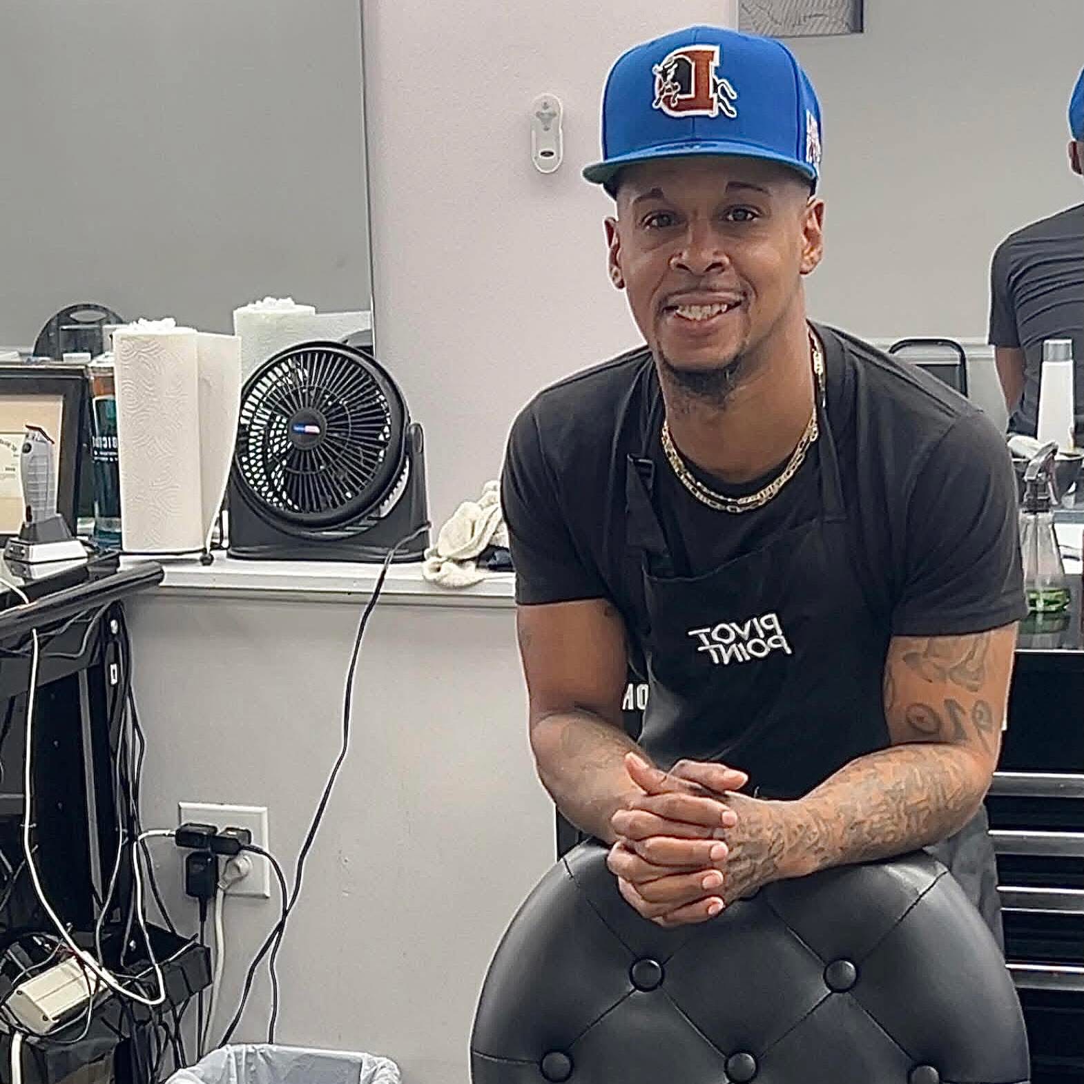 Young man smiling, wearing a blue cap and black t-shirt, sitting in a room with electronic equipment.