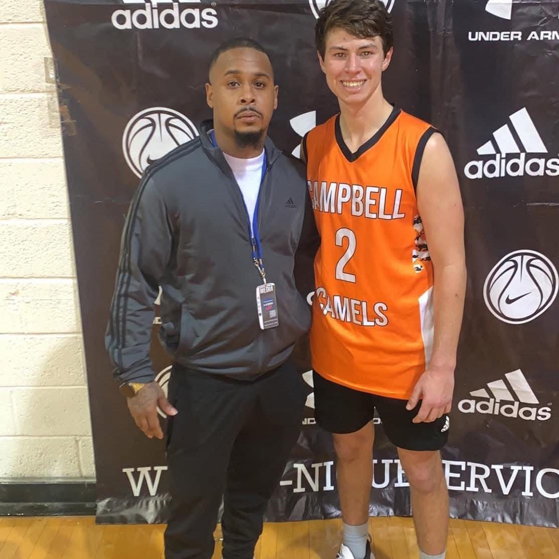 Two young men standing side by side in front of a black backdrop with sports logos, one in a basketball uniform, smiling.