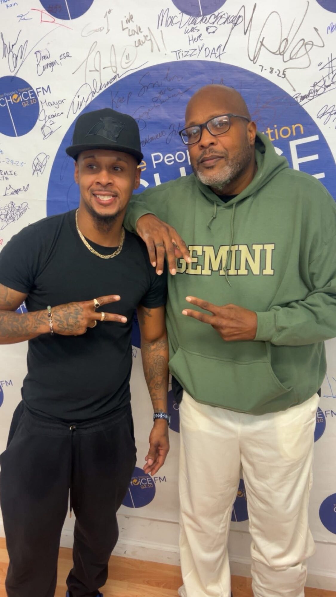 Two men standing together, one wearing a black cap and shirt, the other in a green hoodie, both smiling and making peace signs.