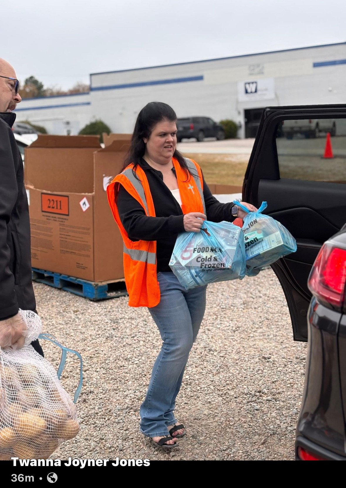 Woman in orange safety vest loading supplies into car trunk, man nearby, outdoor setting with boxes and building in background.