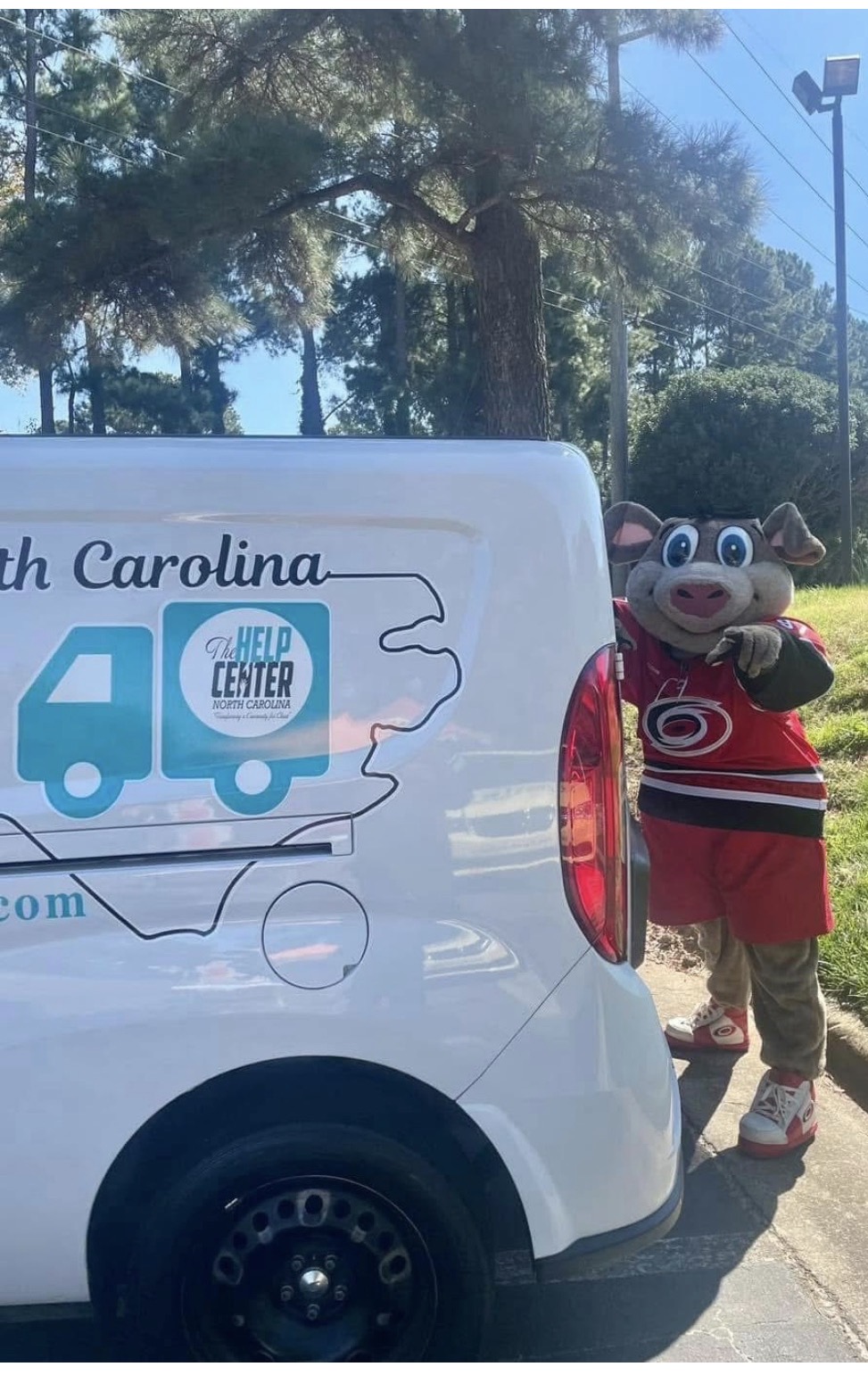 Person in a mascot costume with glasses and a red and black outfit standing outdoors next to a white vehicle with South Carolina map and help center logo, trees in background.