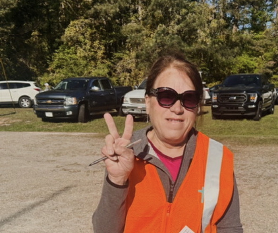Woman wearing sunglasses and an orange safety vest making a peace sign outdoors with trees and parked cars behind her.